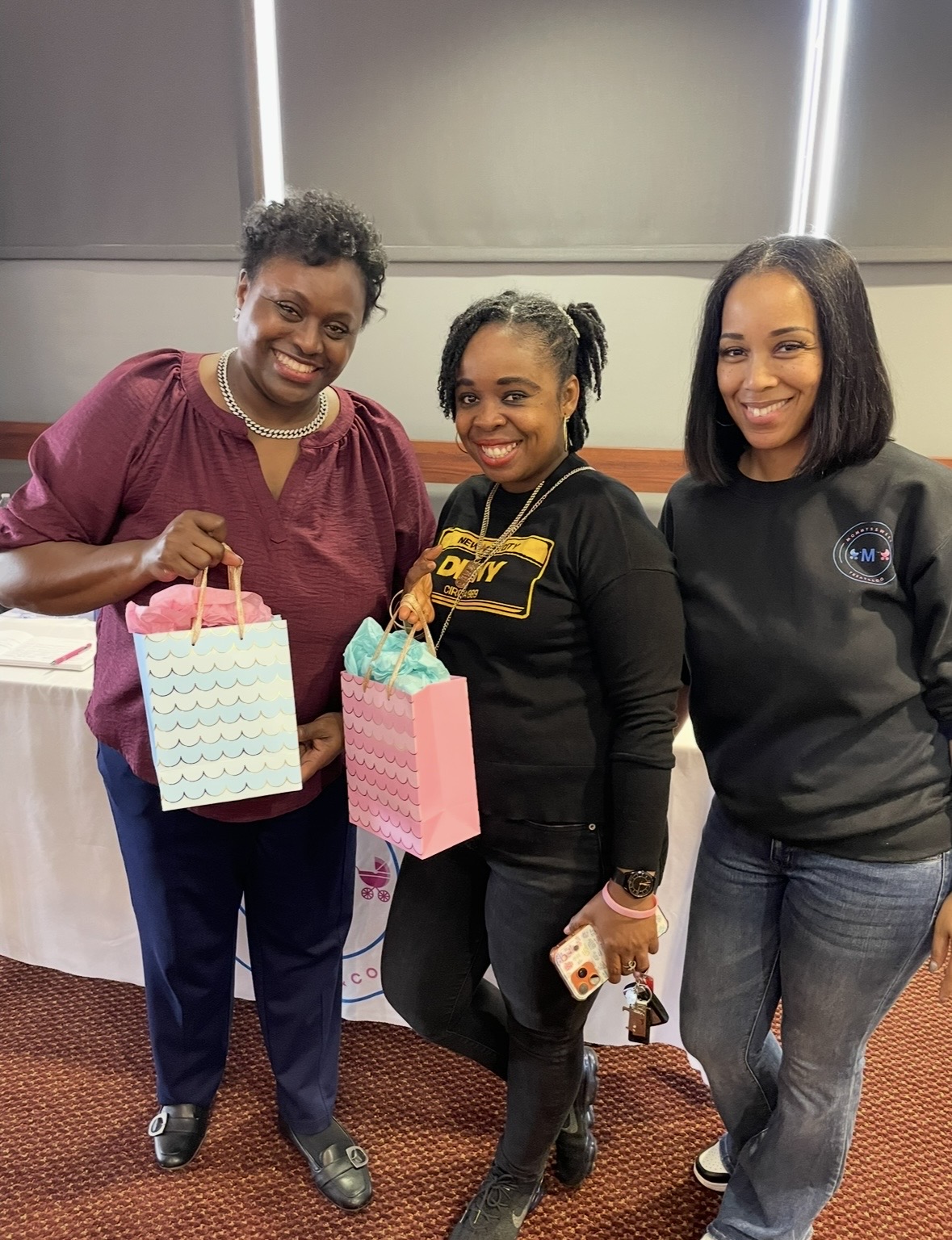 Three women standing indoors, smiling, holding gift bags, with a table and wall behind them.