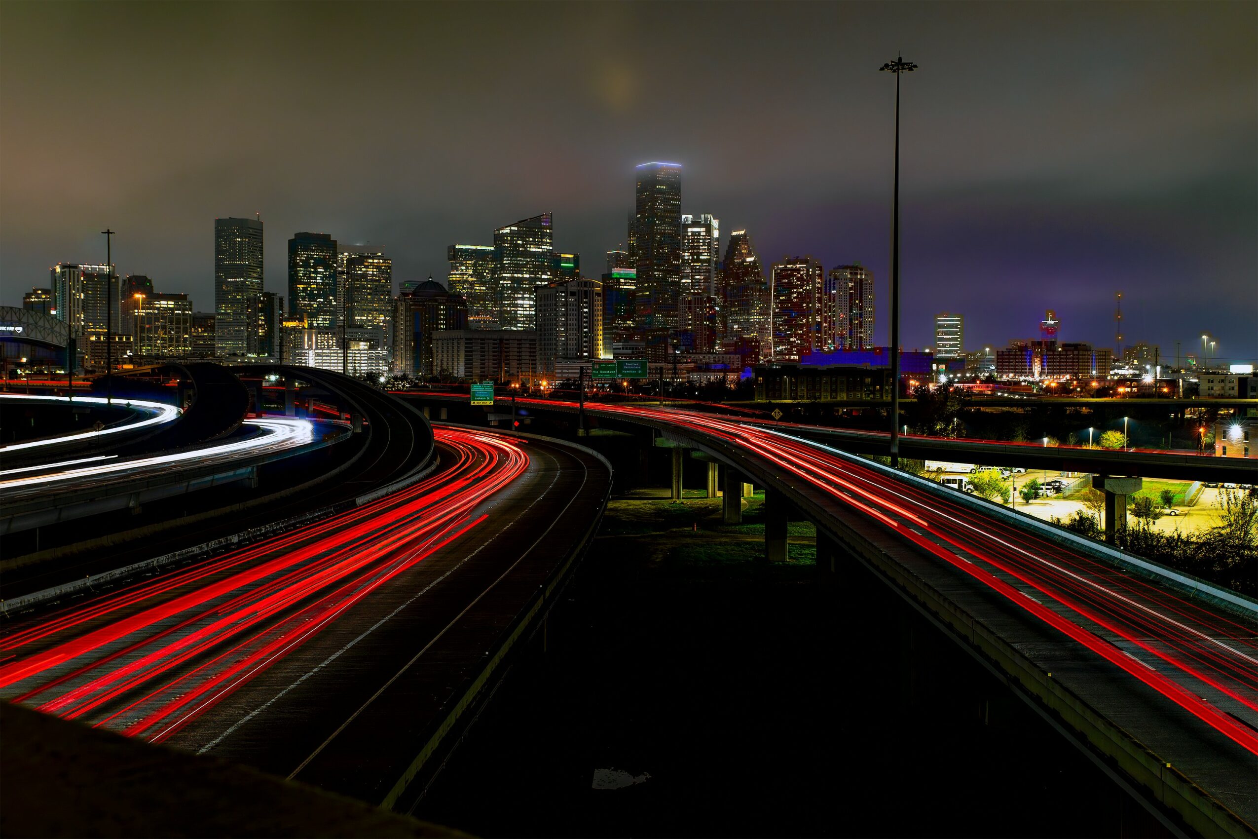 Night city skyline with illuminated buildings and light trails on highway, dark sky, and streetlights.