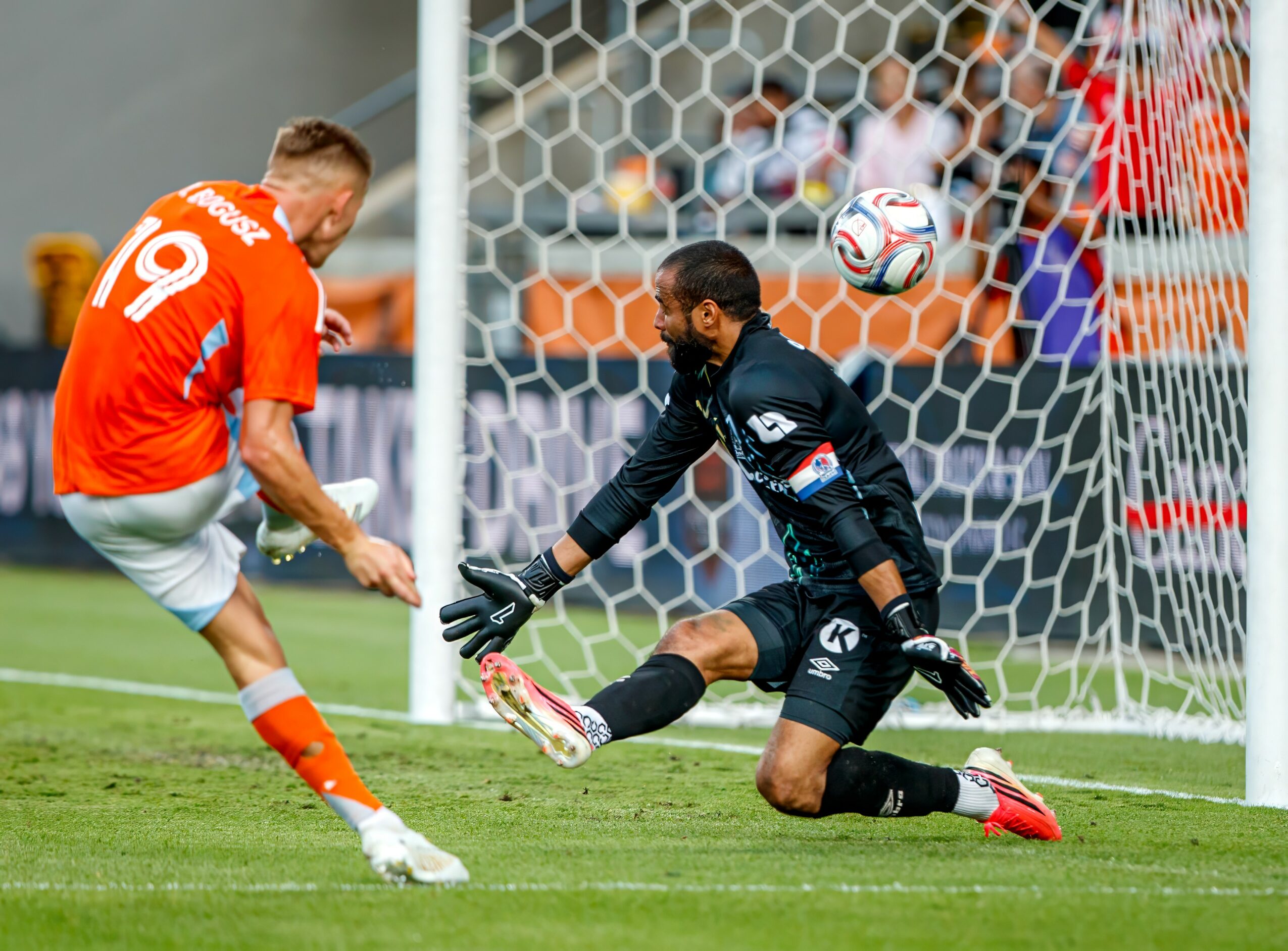 Soccer player in orange kicks ball past goalkeeper in black, near goalpost, with net and spectators in background.