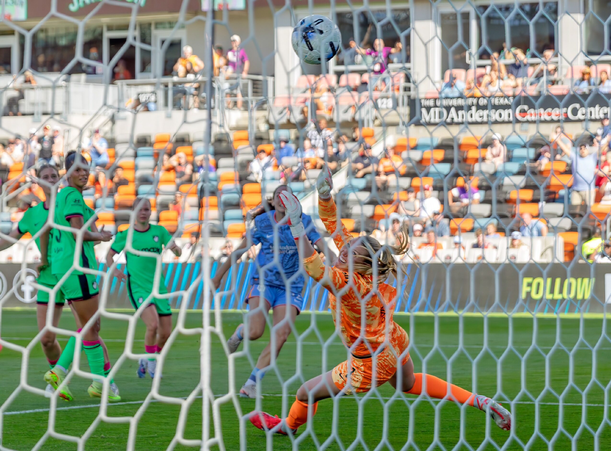 Soccer goalkeeper in orange uniform diving to save a ball during a match, with players and spectators in background.