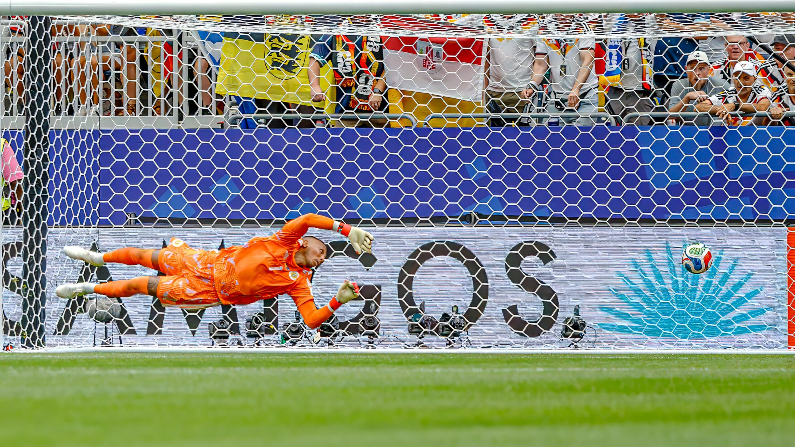 Soccer goalkeeper in orange diving to save a shot, with a net and spectators in the background.