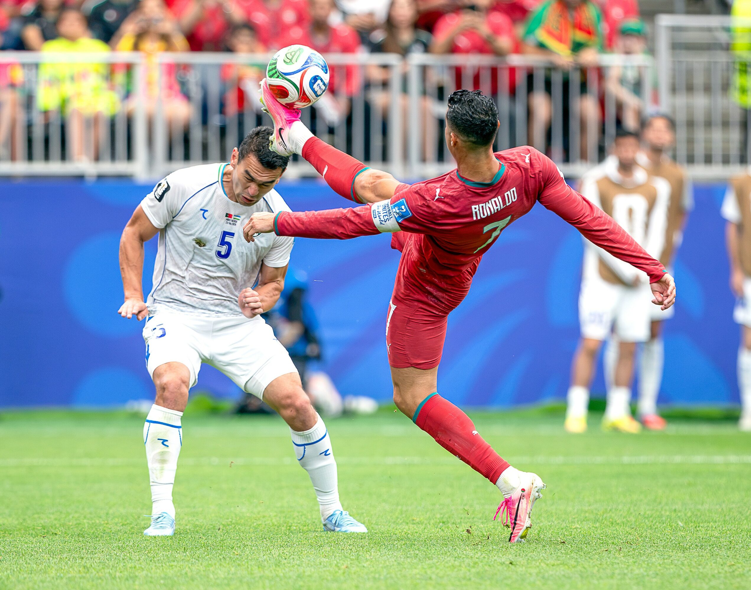 Two soccer players compete for the ball during a match, with one in white and the other in red, on a green field.