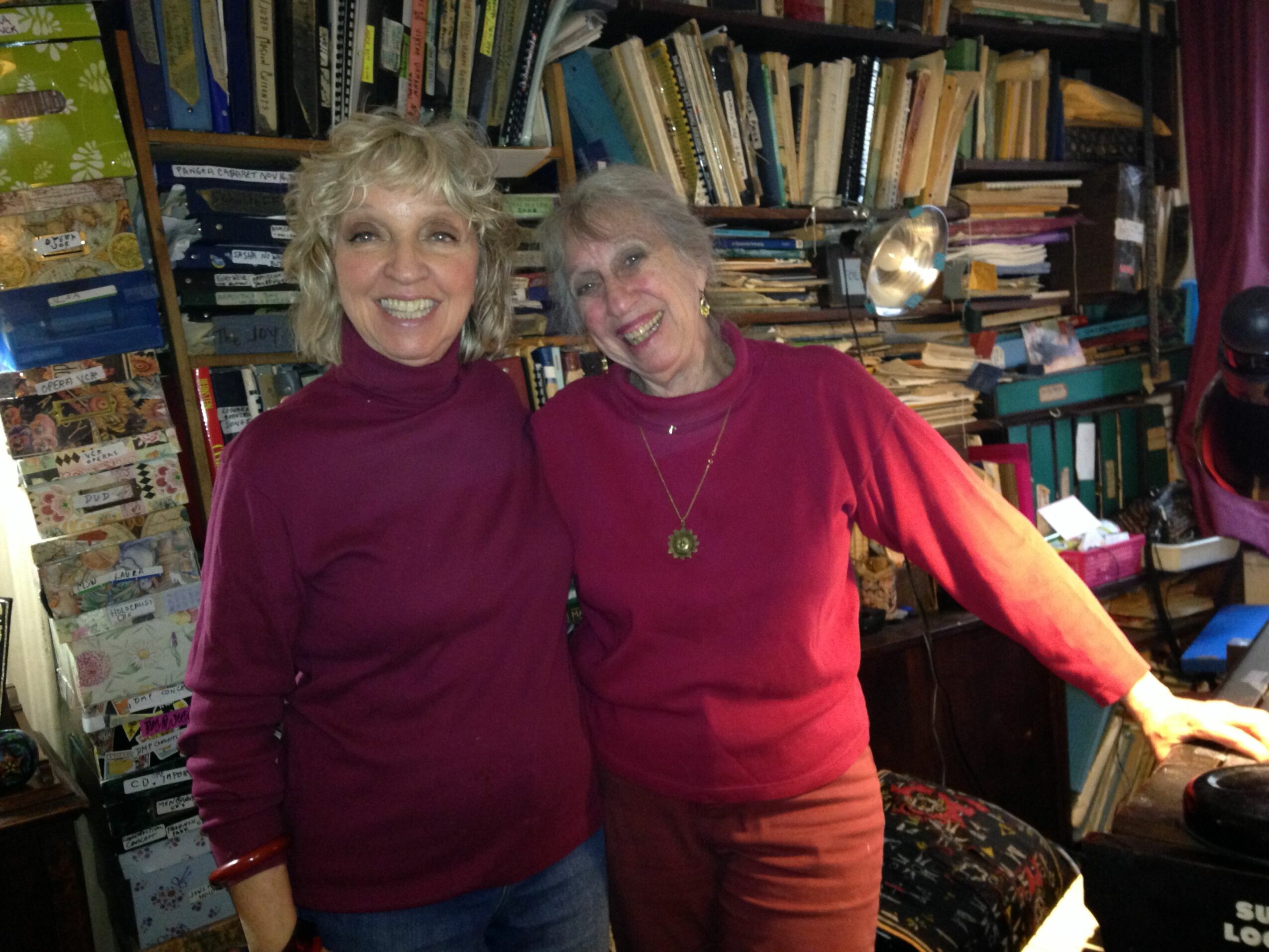Two women with glasses smiling, standing in a bookstore with shelves of books and magazines behind them.