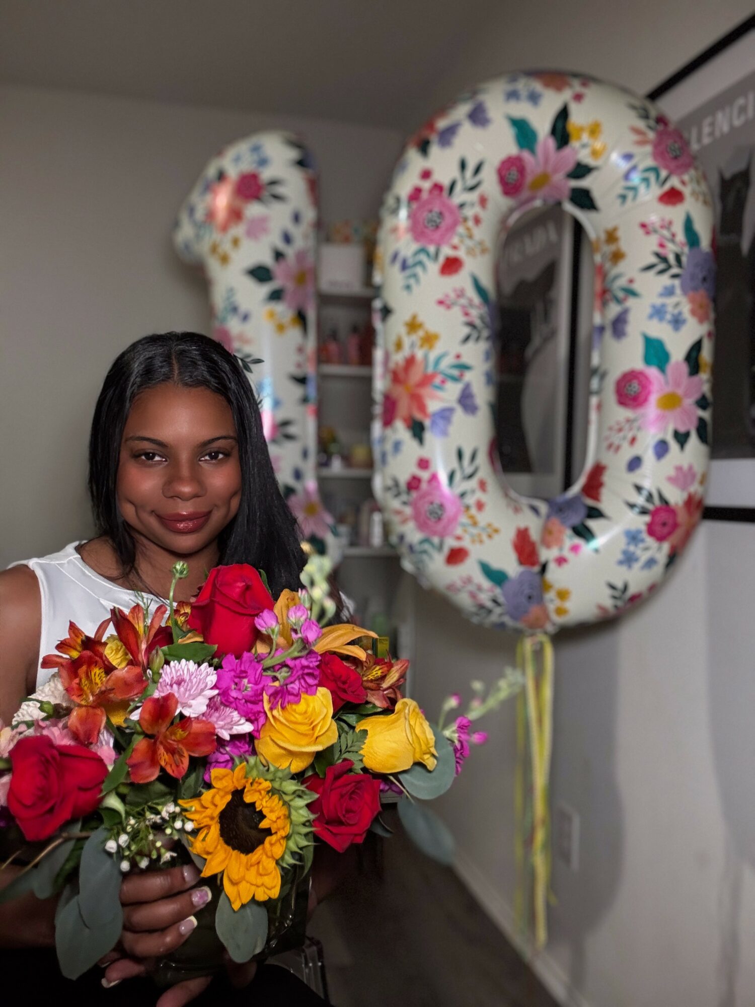 Woman holding a colorful bouquet of flowers in front of large floral number balloons, celebrating an event.