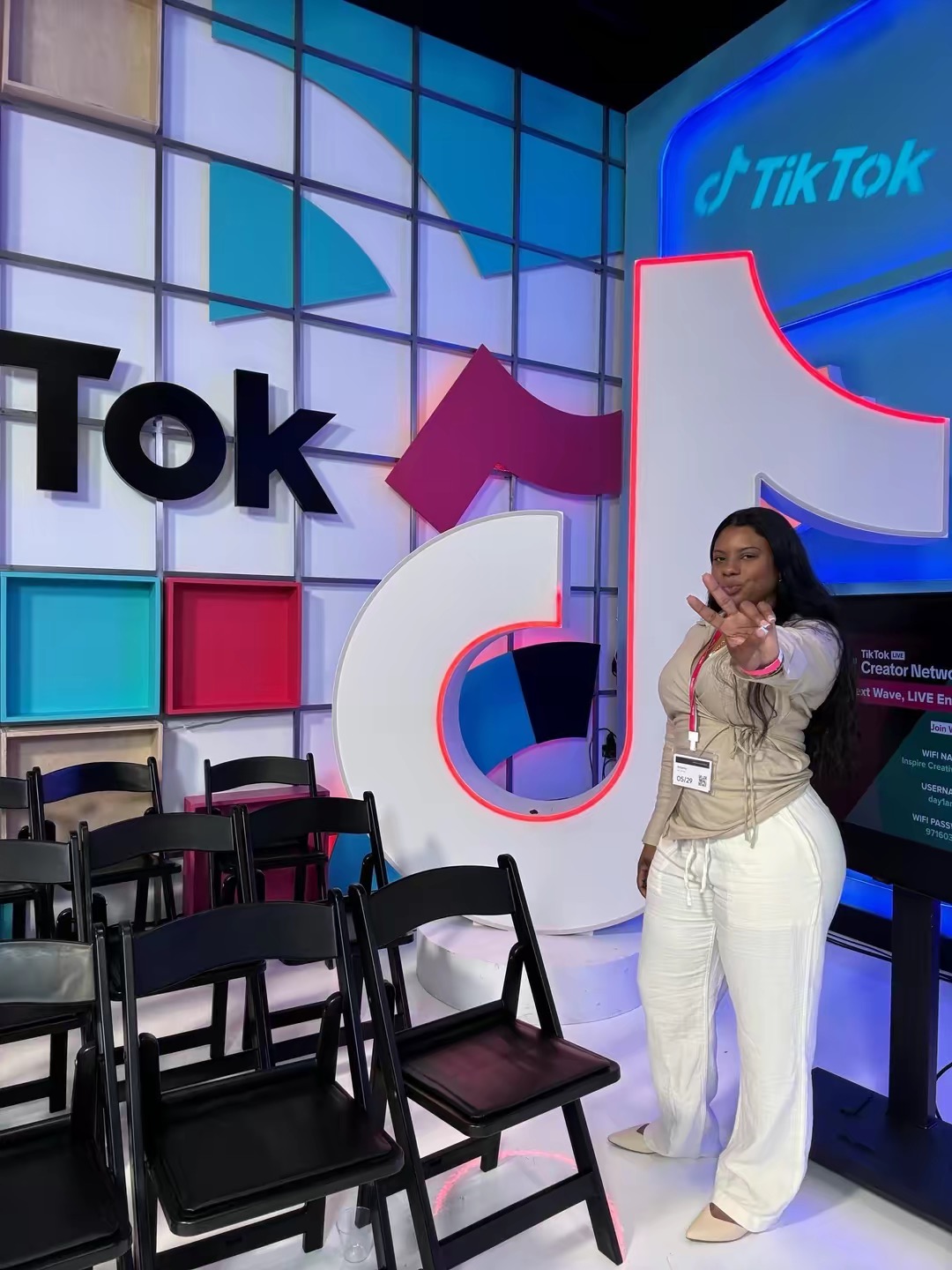 Woman standing in front of TikTok display with chairs, colorful background, and TikTok logo, smiling and pointing.