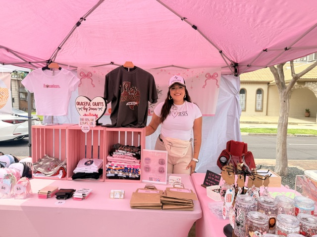 Young woman standing behind a pink booth with clothing and accessories, outdoors on a sunny day.