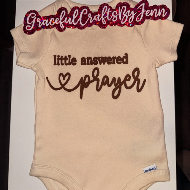 Cream-colored onesie with brown text reading 'little answered prayer' and a heart shape, with a pink and red sign on top that says 'Graceful Crafts By Jenn'.