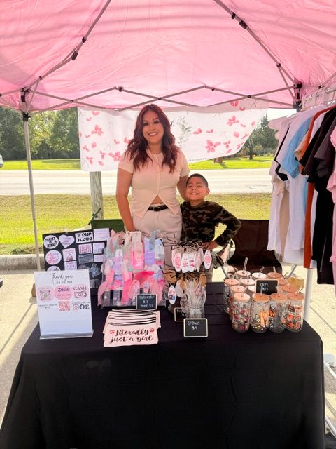 Woman standing behind a table with pink and clear bottles, signs, and displays outdoors, with clothing hanging nearby.