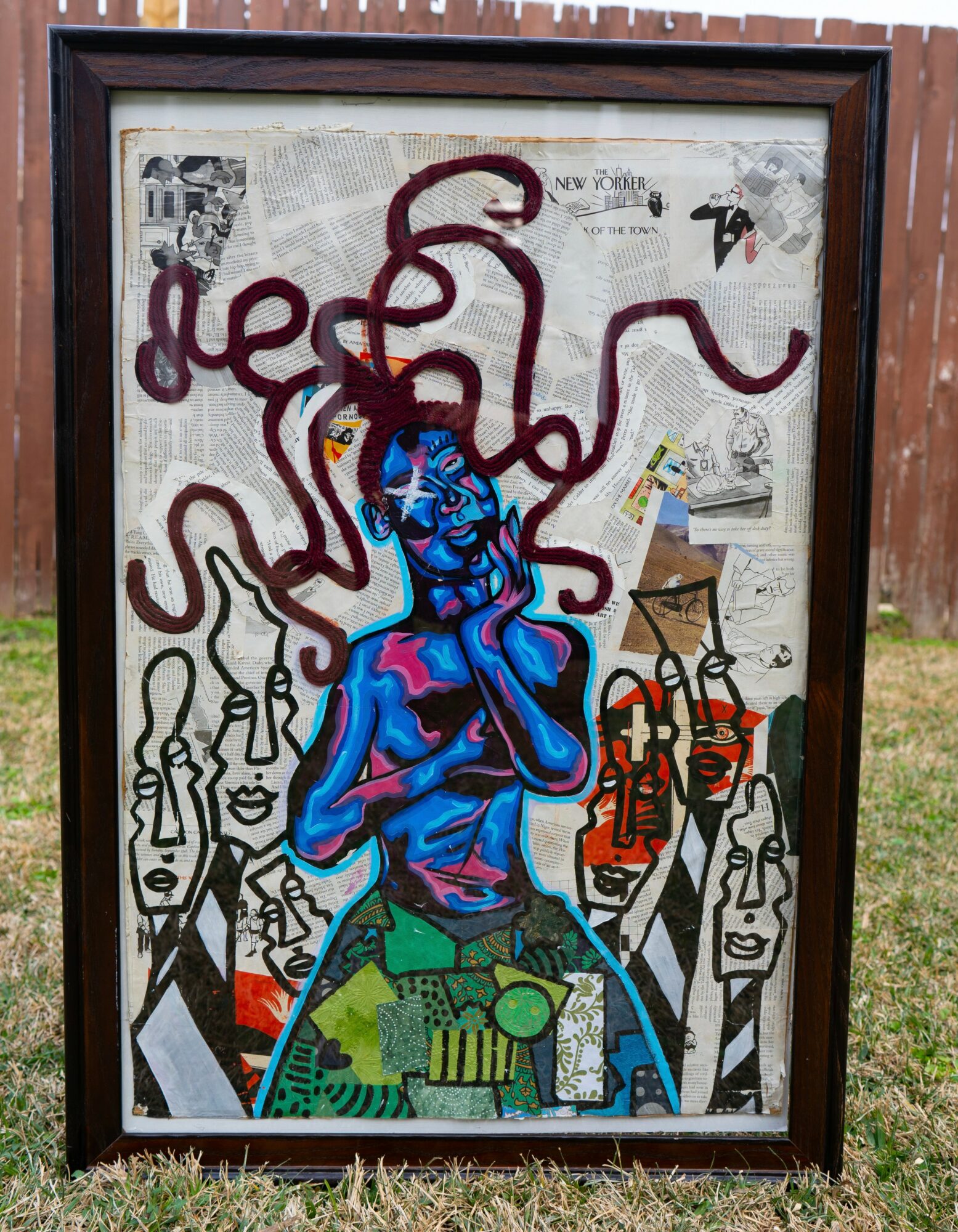 Framed street art mural featuring a blue figure with a red snake-like shape on head, surrounded by black and white faces, and colorful objects.