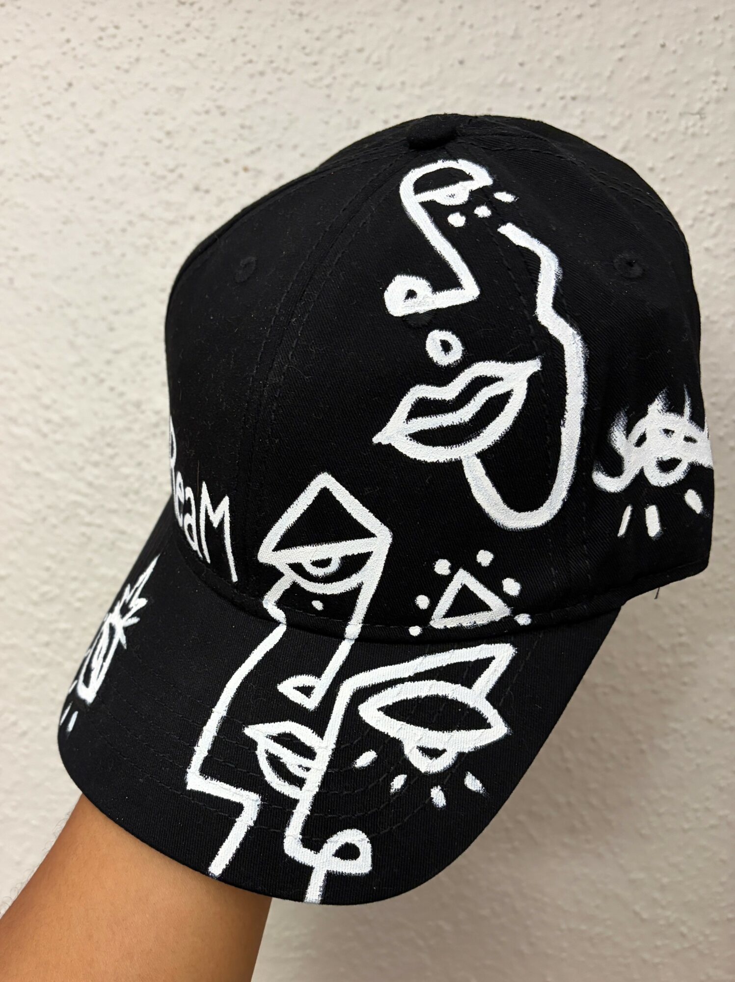 Black baseball cap with white abstract face and geometric designs, worn by a person against a light background.