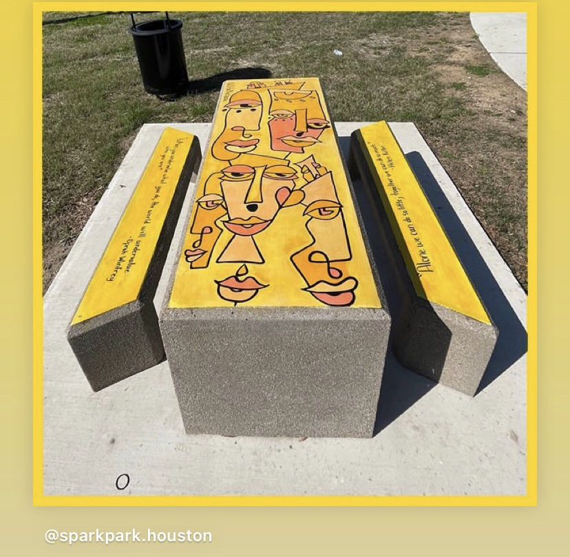 Concrete bench with yellow painted abstract faces, two yellow armrests, and a black trash can in the background, outdoors.