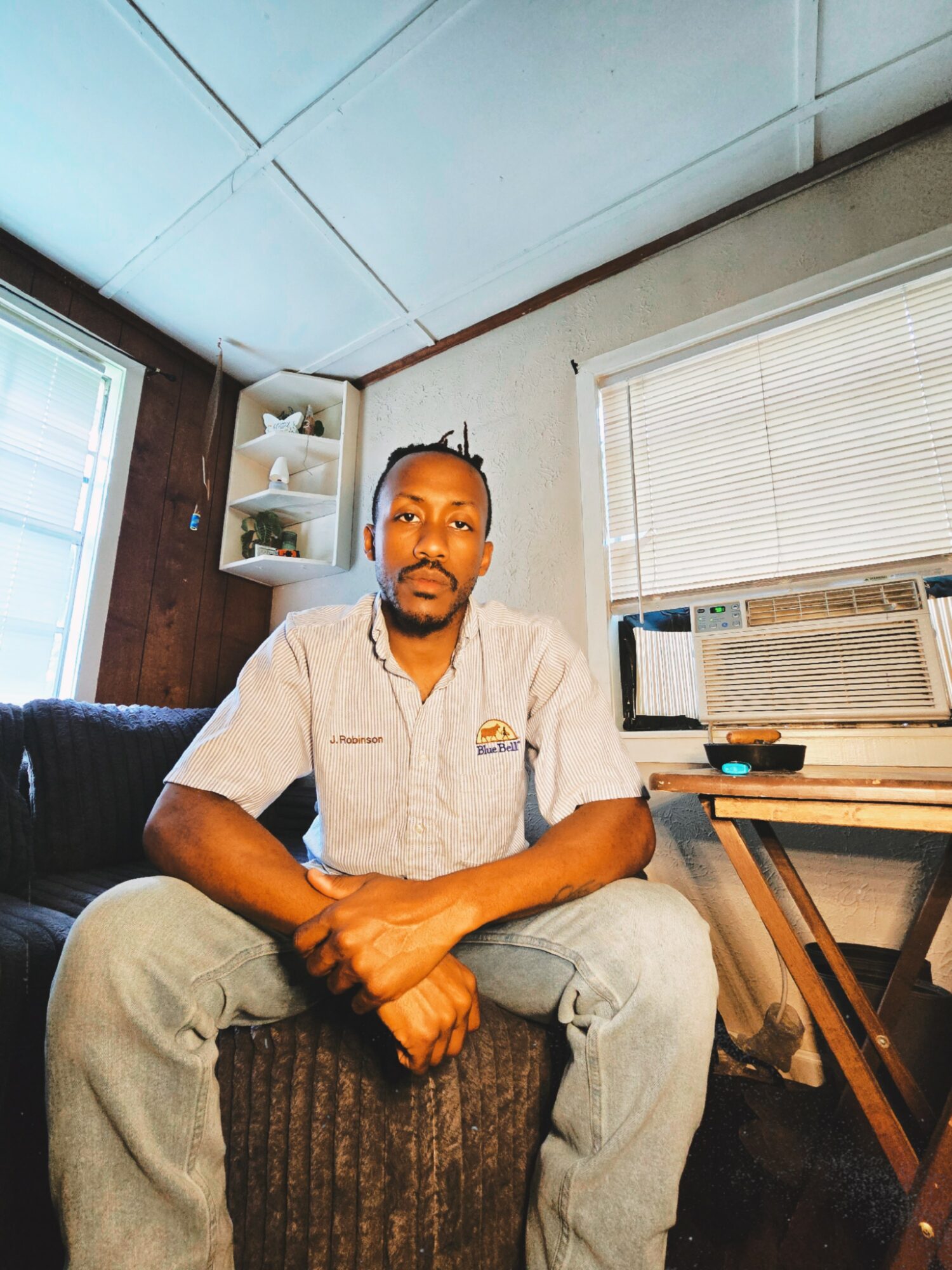 Man sitting on a couch with arms crossed, in a room with a window air conditioner and blinds, wooden wall, and shelves.