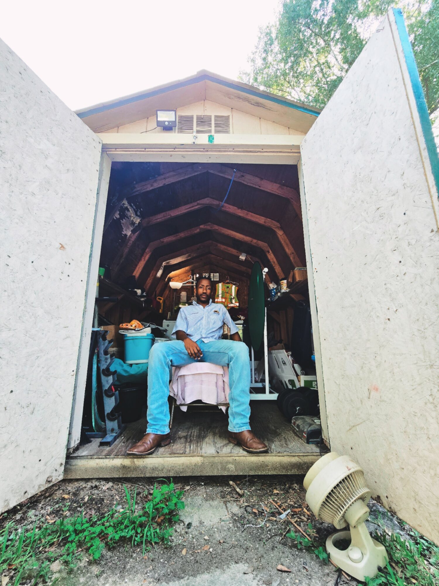 Person sitting on a bed inside a small open structure, with open doors and a house in the background.