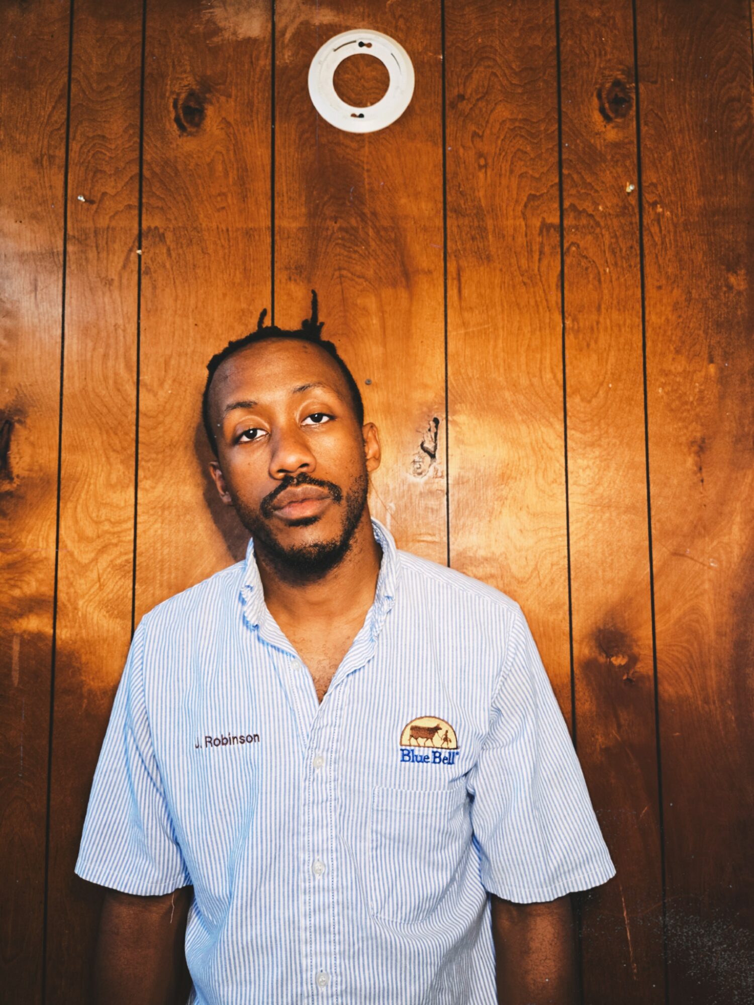 Man with a beard and mustache wearing a light blue uniform shirt standing against a wooden wall.