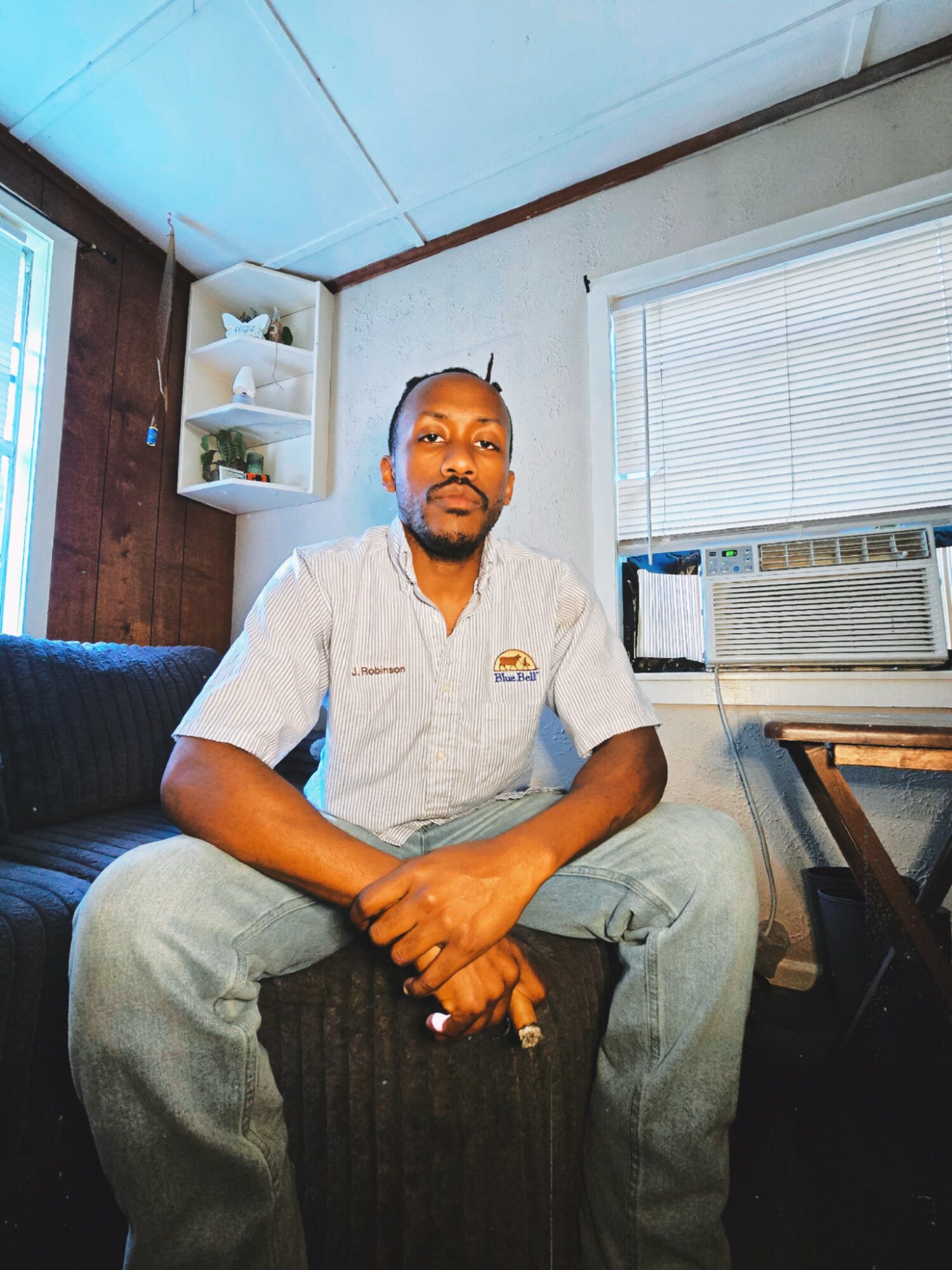 Man sitting on a black ottoman in a room with white walls, window with blinds, and an air conditioning unit.