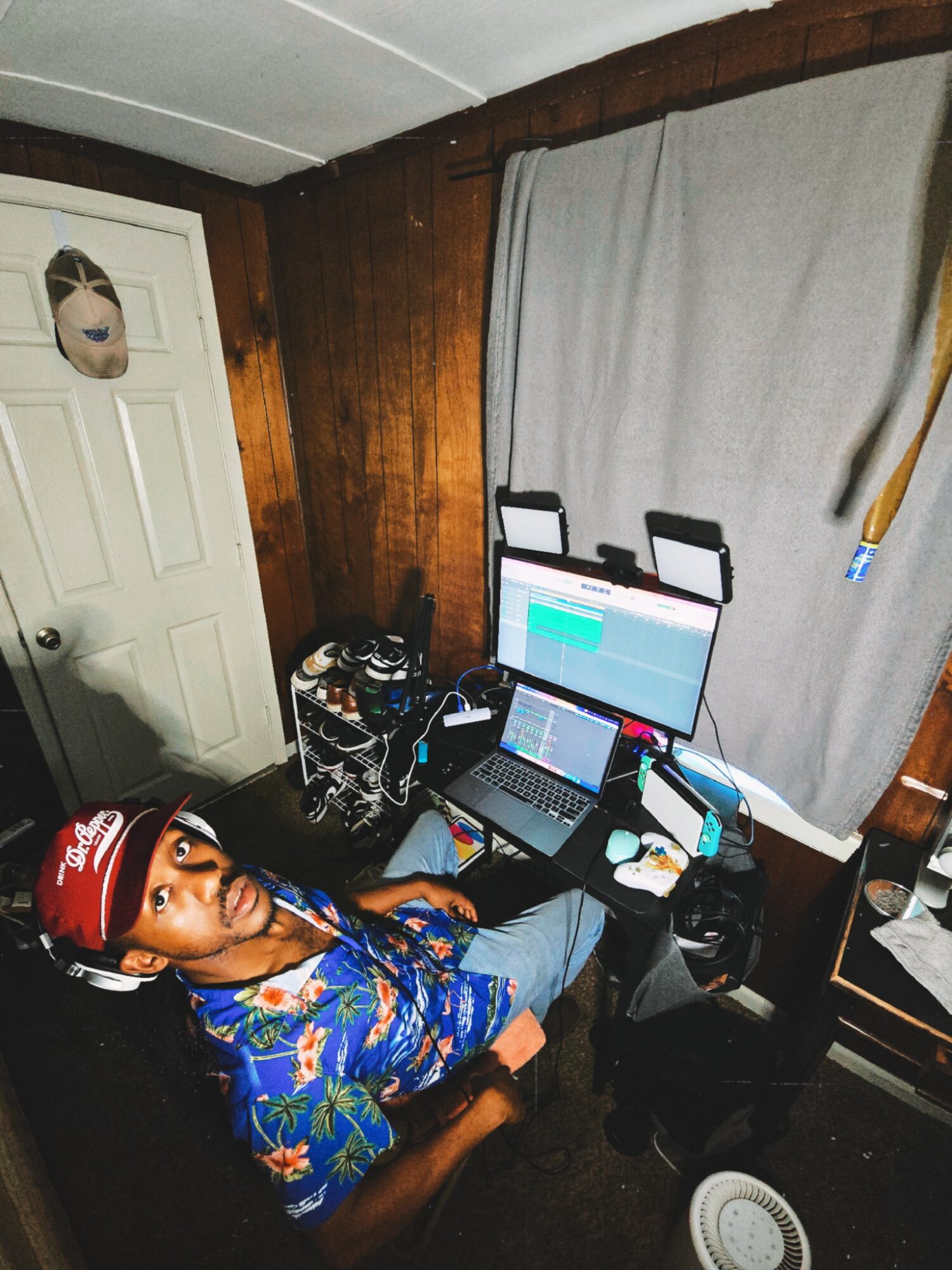 Young person wearing a red cap and colorful shirt sitting at a cluttered desk with multiple electronic devices, in a room with wood-paneled walls and a window with curtains.