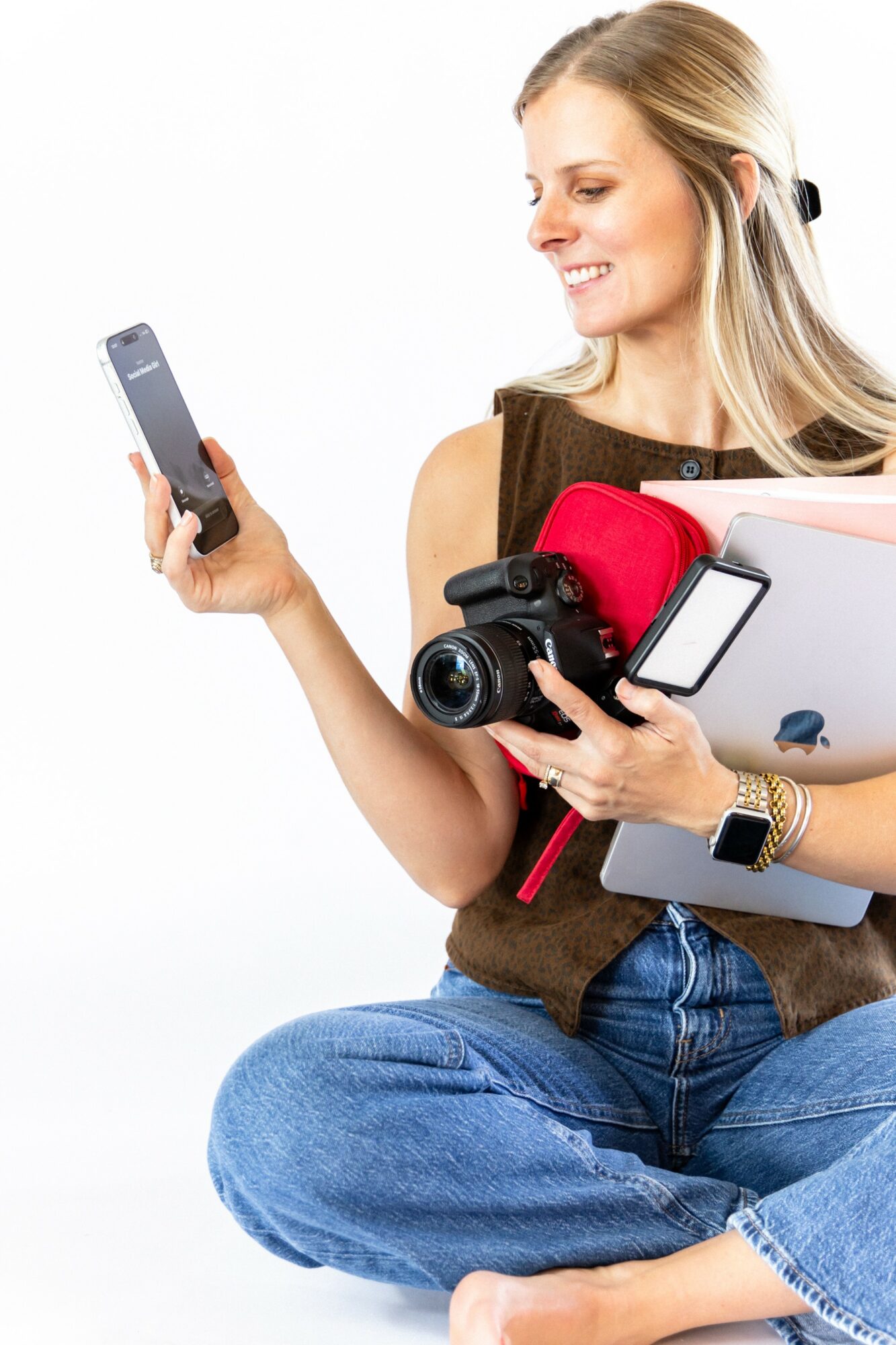 Woman sitting cross-legged, holding a smartphone, camera, and a tablet, smiling and looking at her phone.