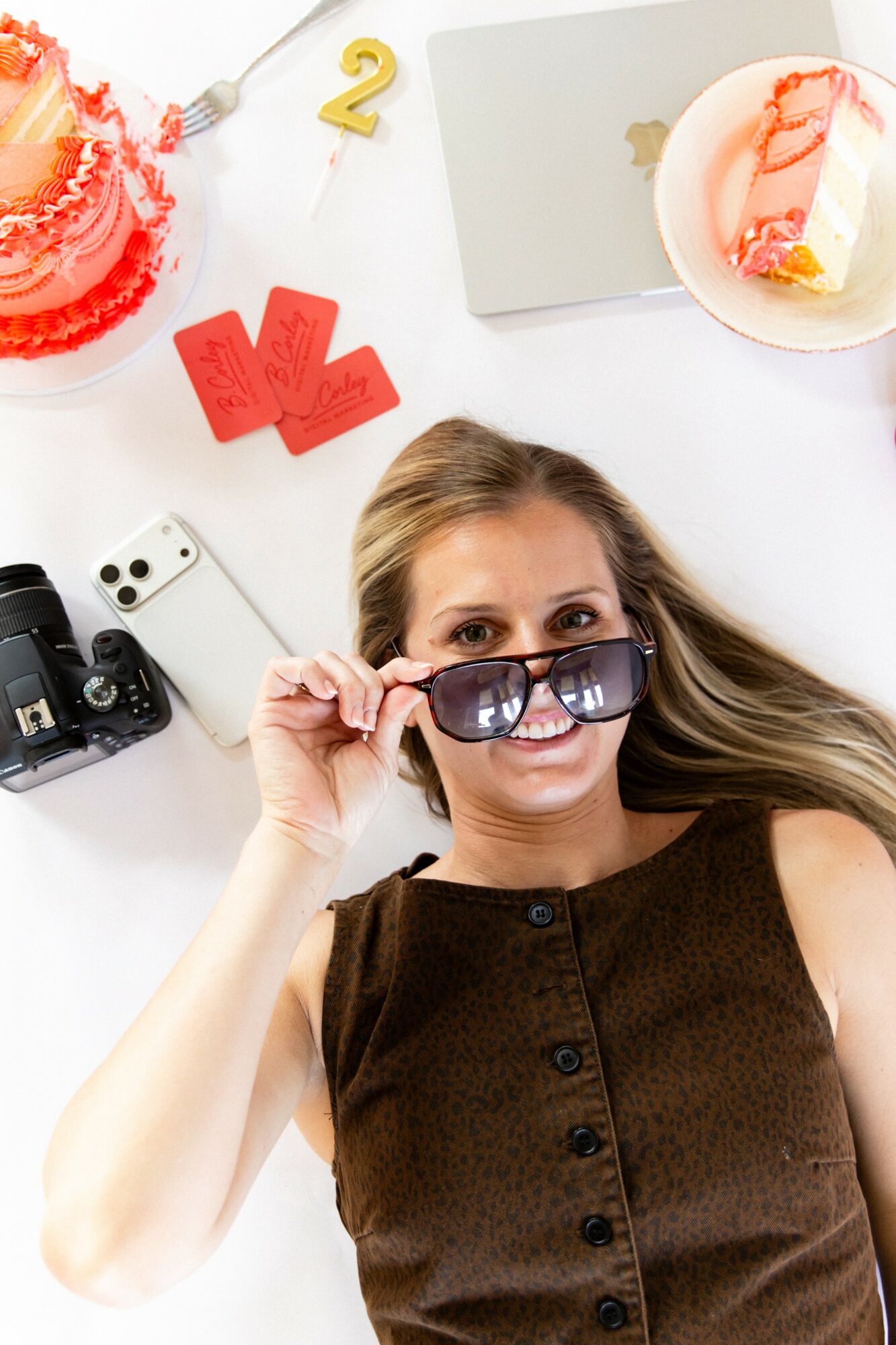 Woman with long hair and sunglasses lying on a white surface surrounded by cake, camera, and birthday decorations.
