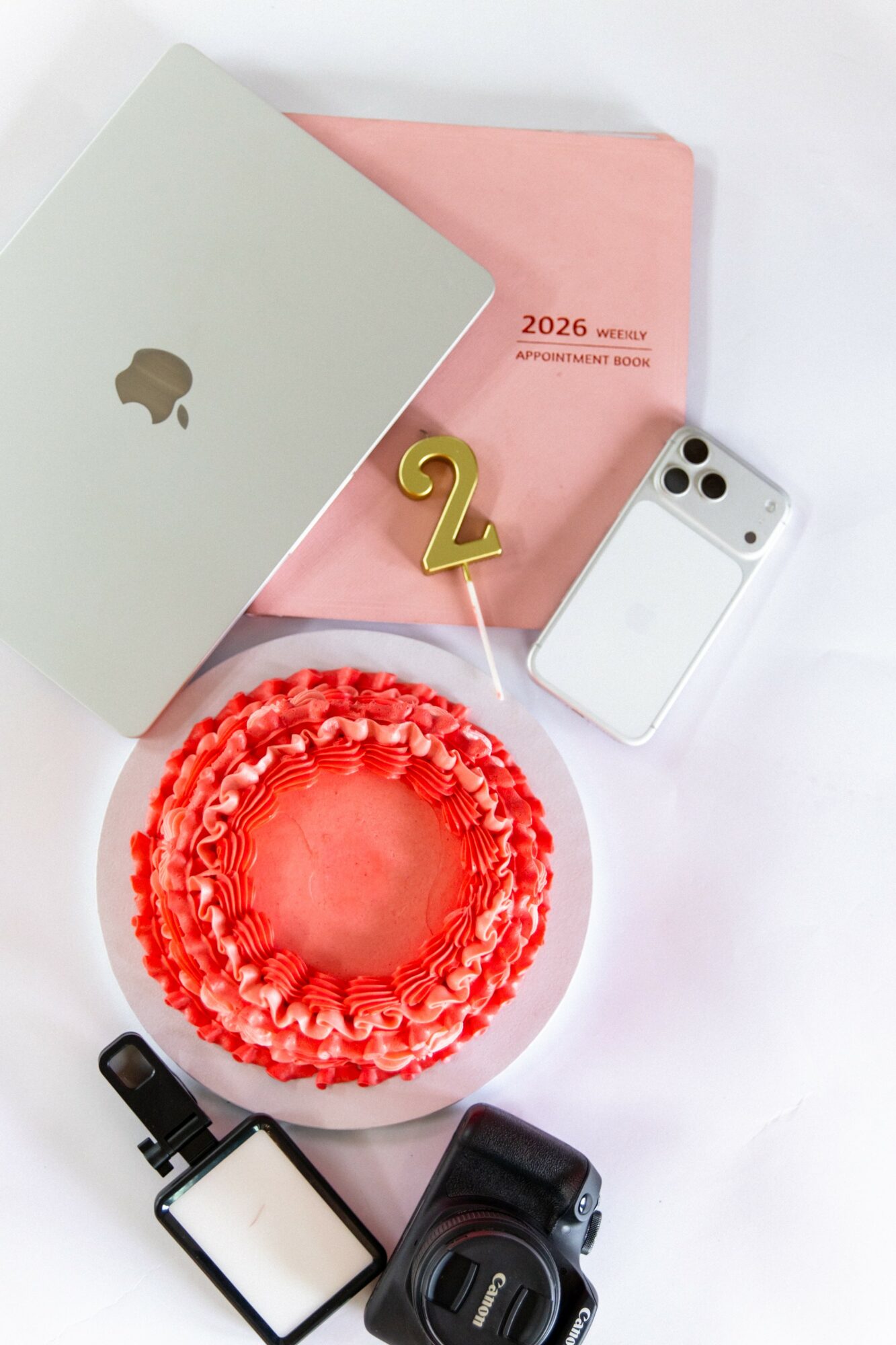 Flat lay of a silver MacBook, pink notebook, white iPhone, red cake, camera, and clip on a white surface.