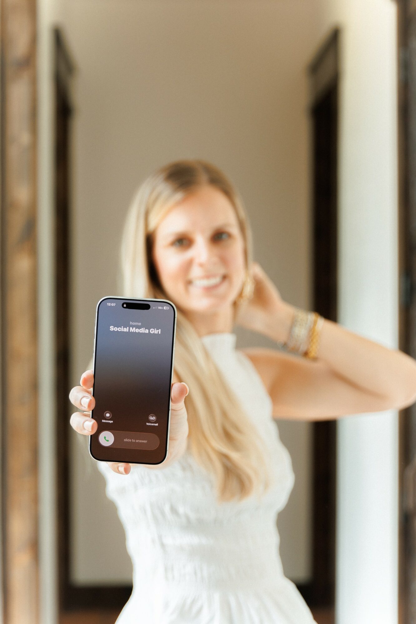 Woman with long blonde hair smiling and holding a smartphone towards the camera in a hallway.