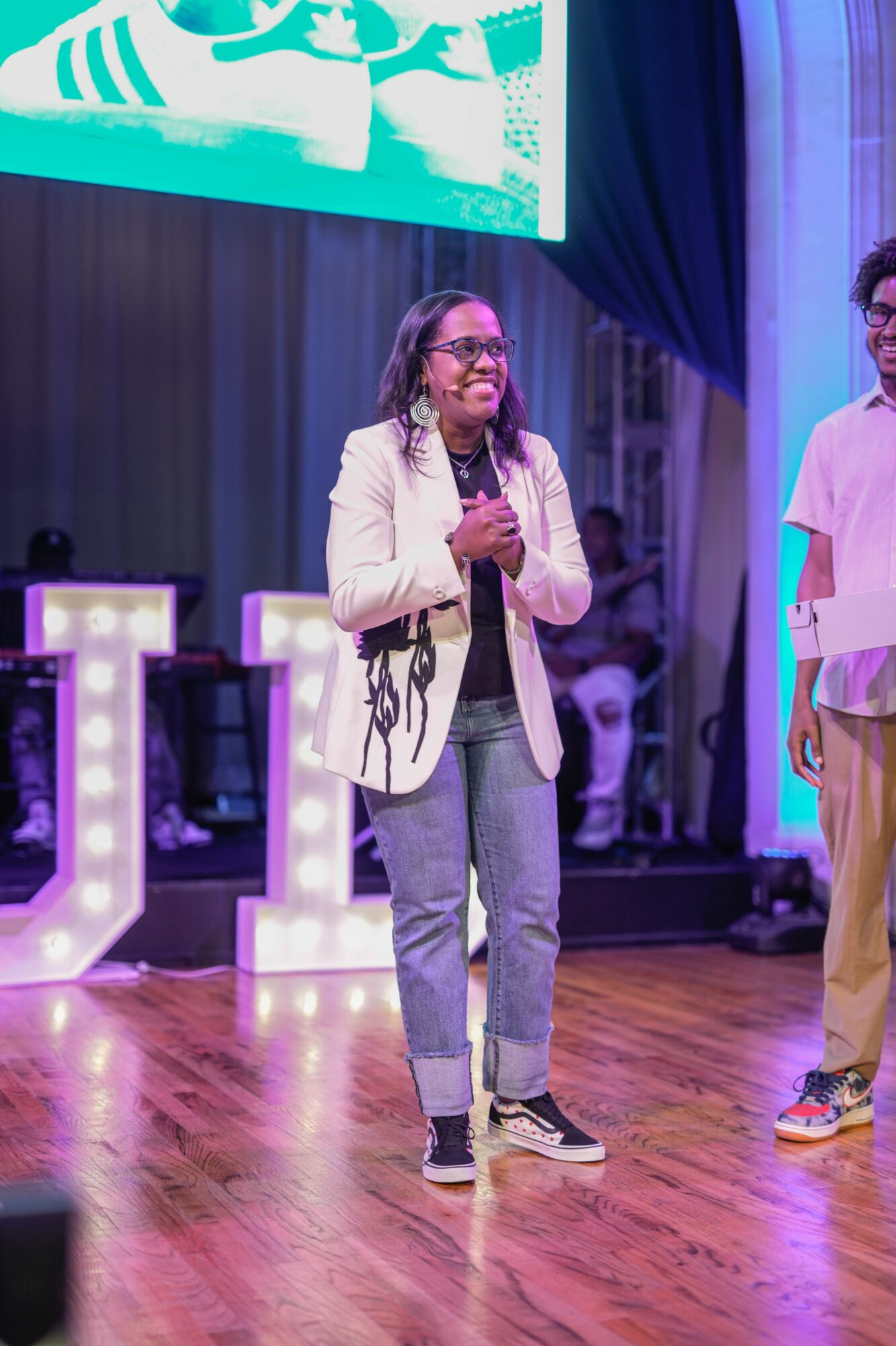 Woman speaking on stage with a microphone, wearing glasses, white blazer, jeans, and sneakers, in a decorated event space.