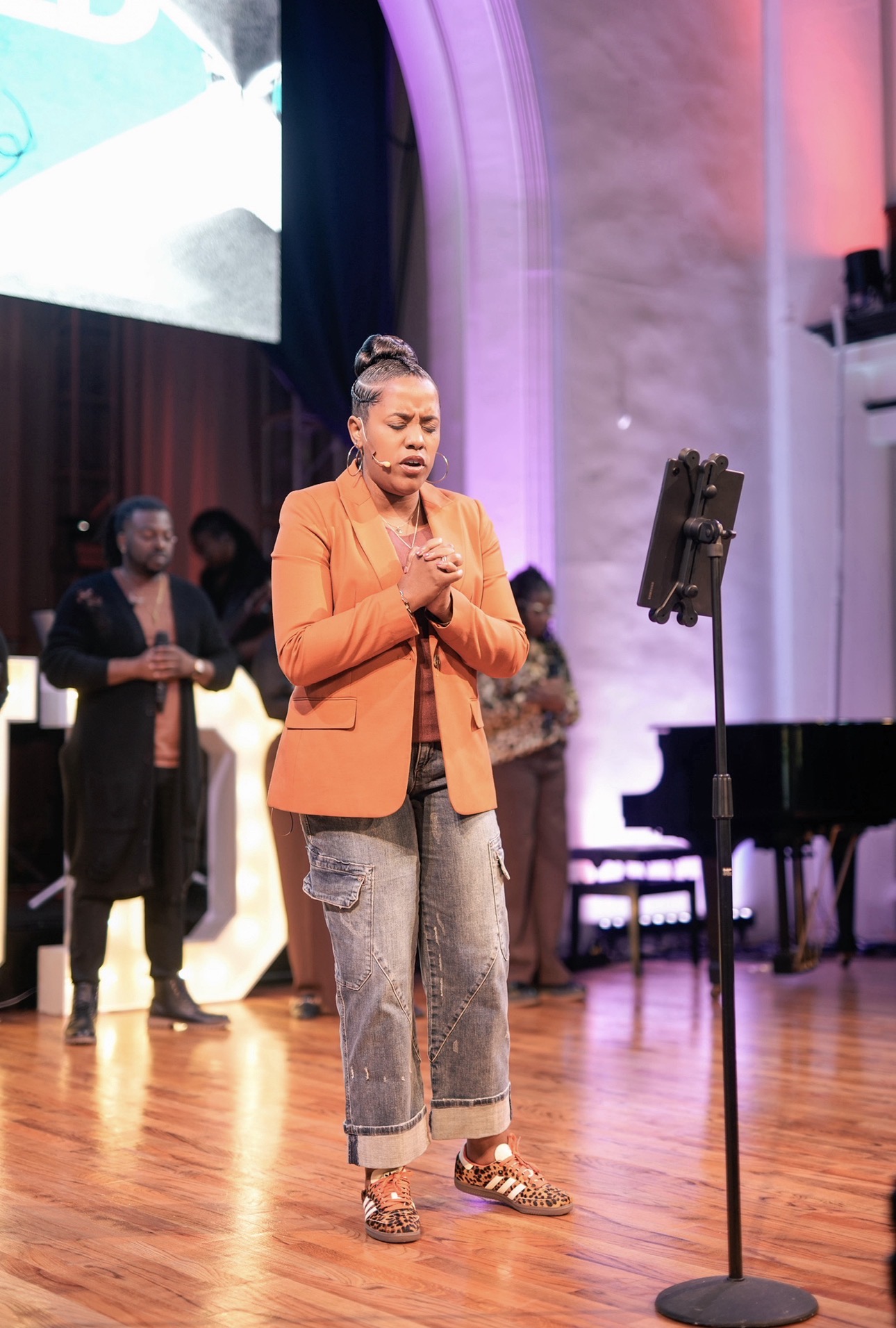 Woman in orange blazer and jeans praying on stage with people and a piano in background.