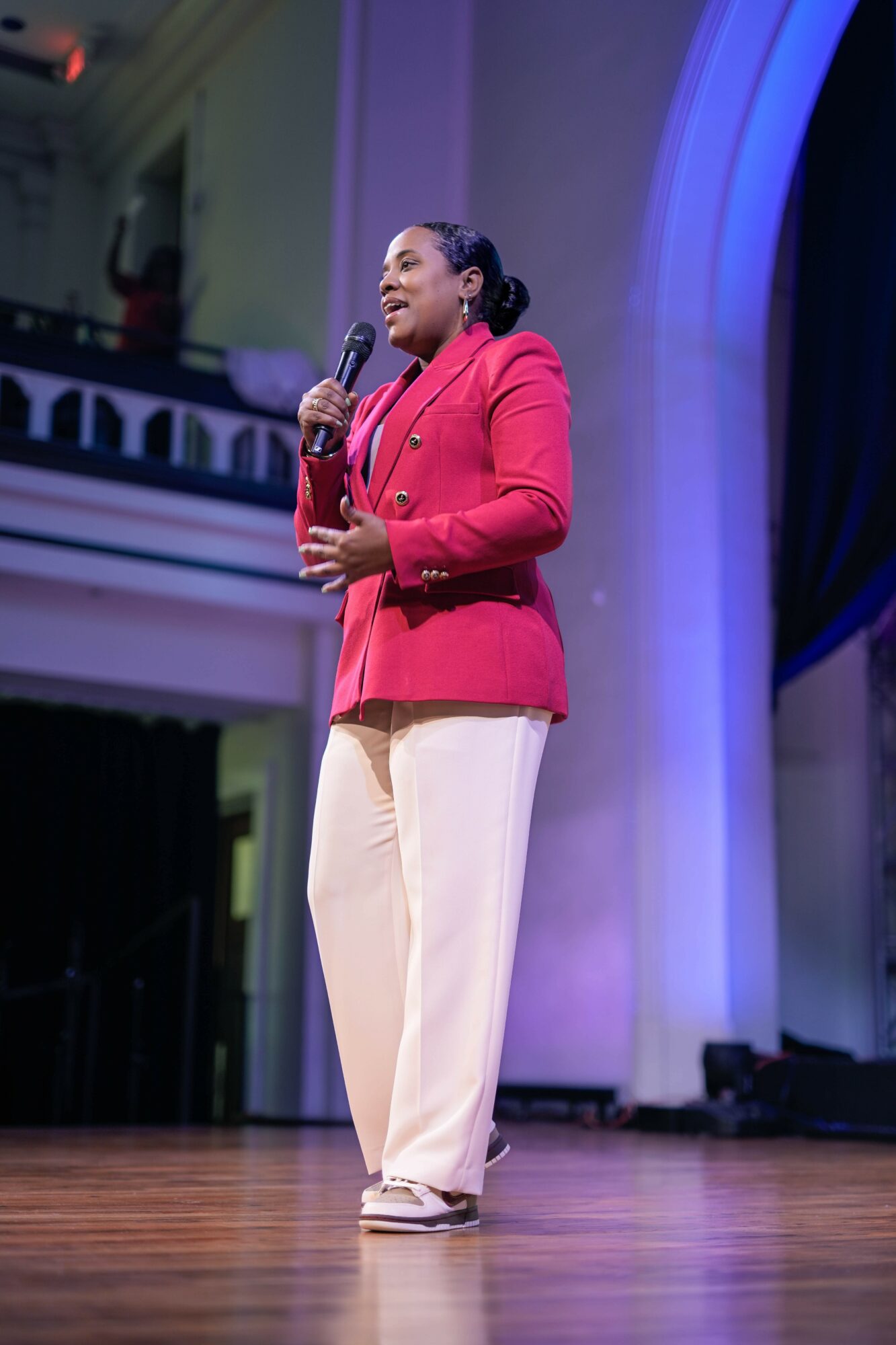 Woman in red blazer and beige pants speaking into a microphone on stage, with a blue-lit arch in the background.