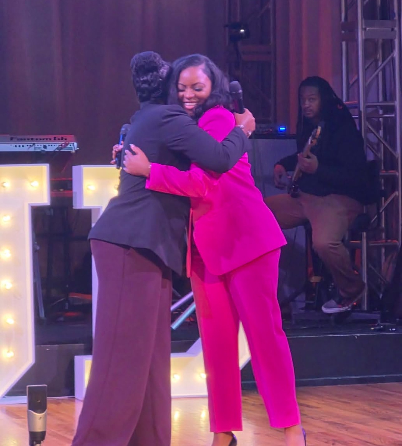 Two women hugging on stage, one in a pink suit, the other in dark clothing, with a musician in the background.