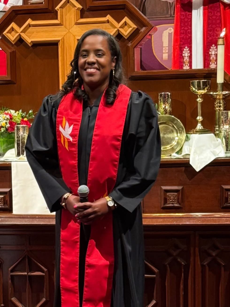 Woman in graduation gown and red stole holding a microphone, standing in front of an altar with candles and religious symbols.