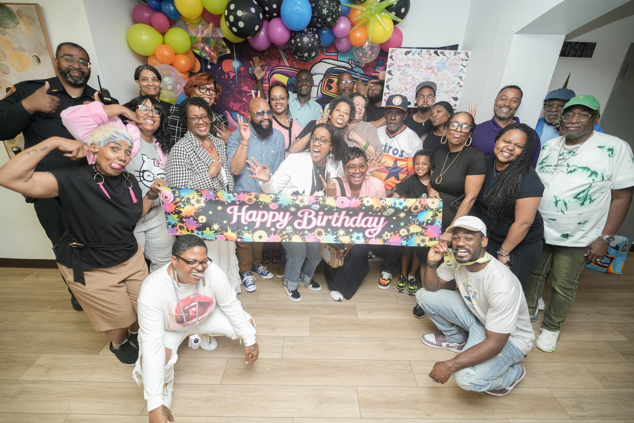 Group of people celebrating birthday with balloons, holding a banner that says 'Happy Birthday,' smiling indoors.