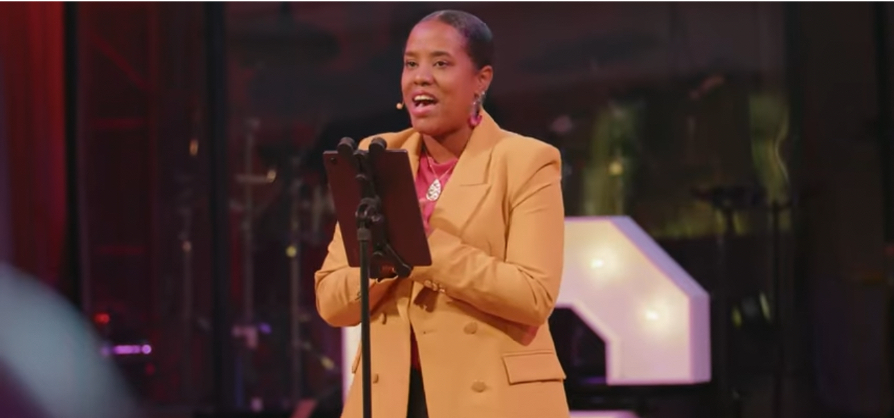 Woman in yellow blazer speaking into microphone on stage, holding a tablet, smiling, with a dark background.