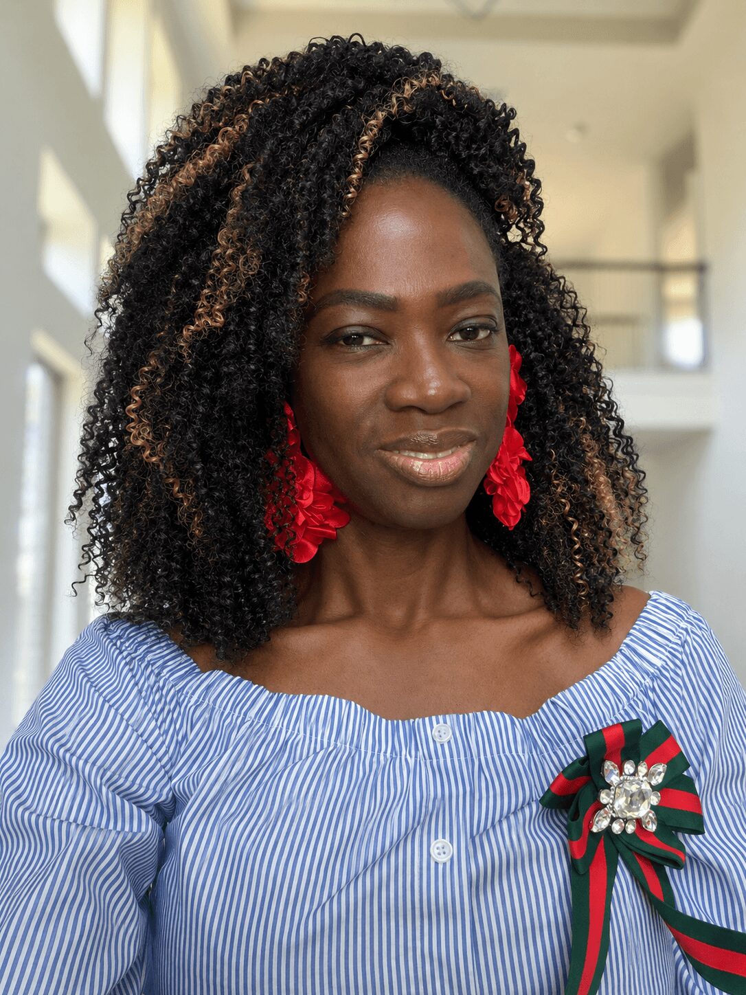 Woman with curly hair wearing a blue and white striped off-shoulder top and red earrings, indoors.