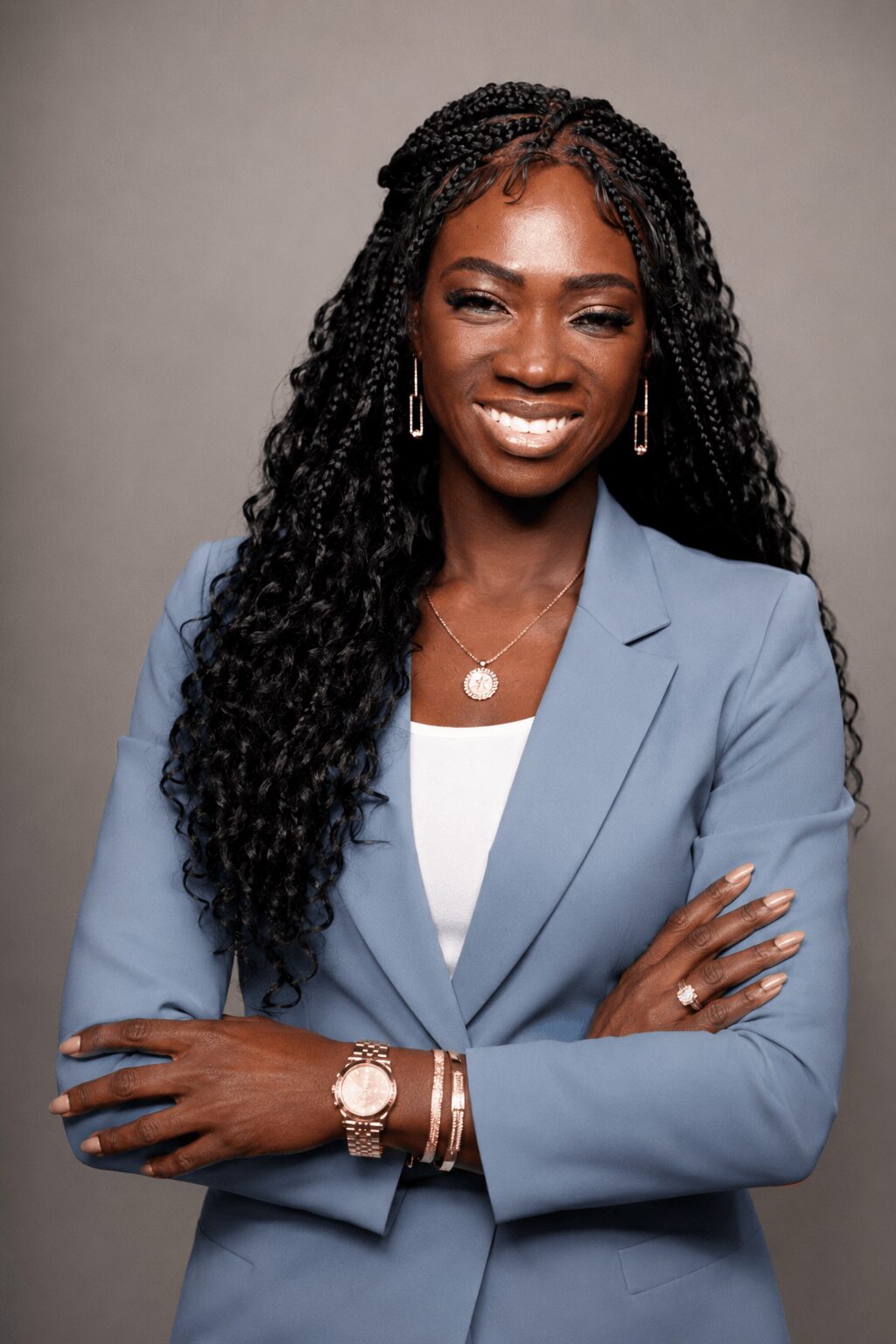 Woman with long curly hair wearing a light blue blazer and jewelry, smiling with arms crossed.