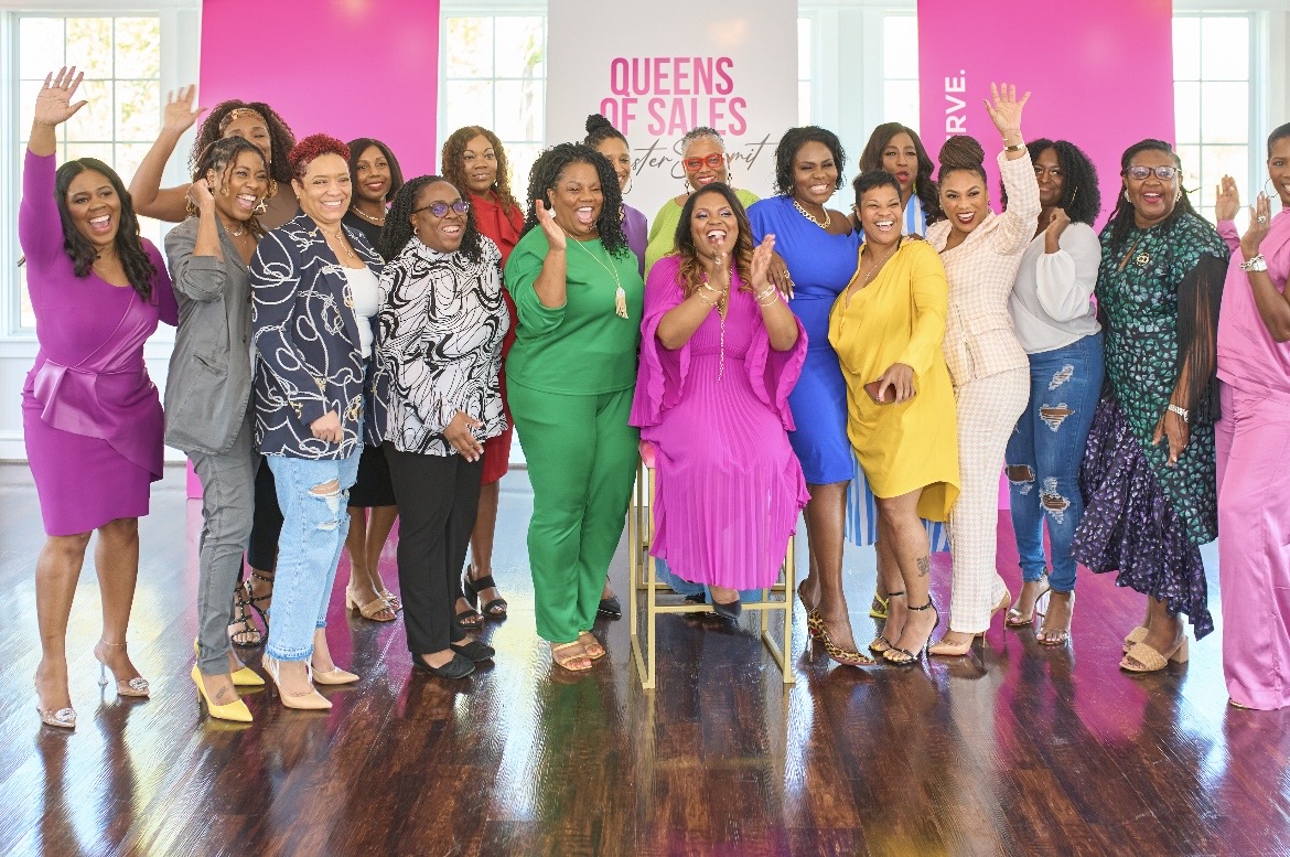 Group of women standing and sitting in a room with pink banners, celebrating a sale event, smiling and waving.