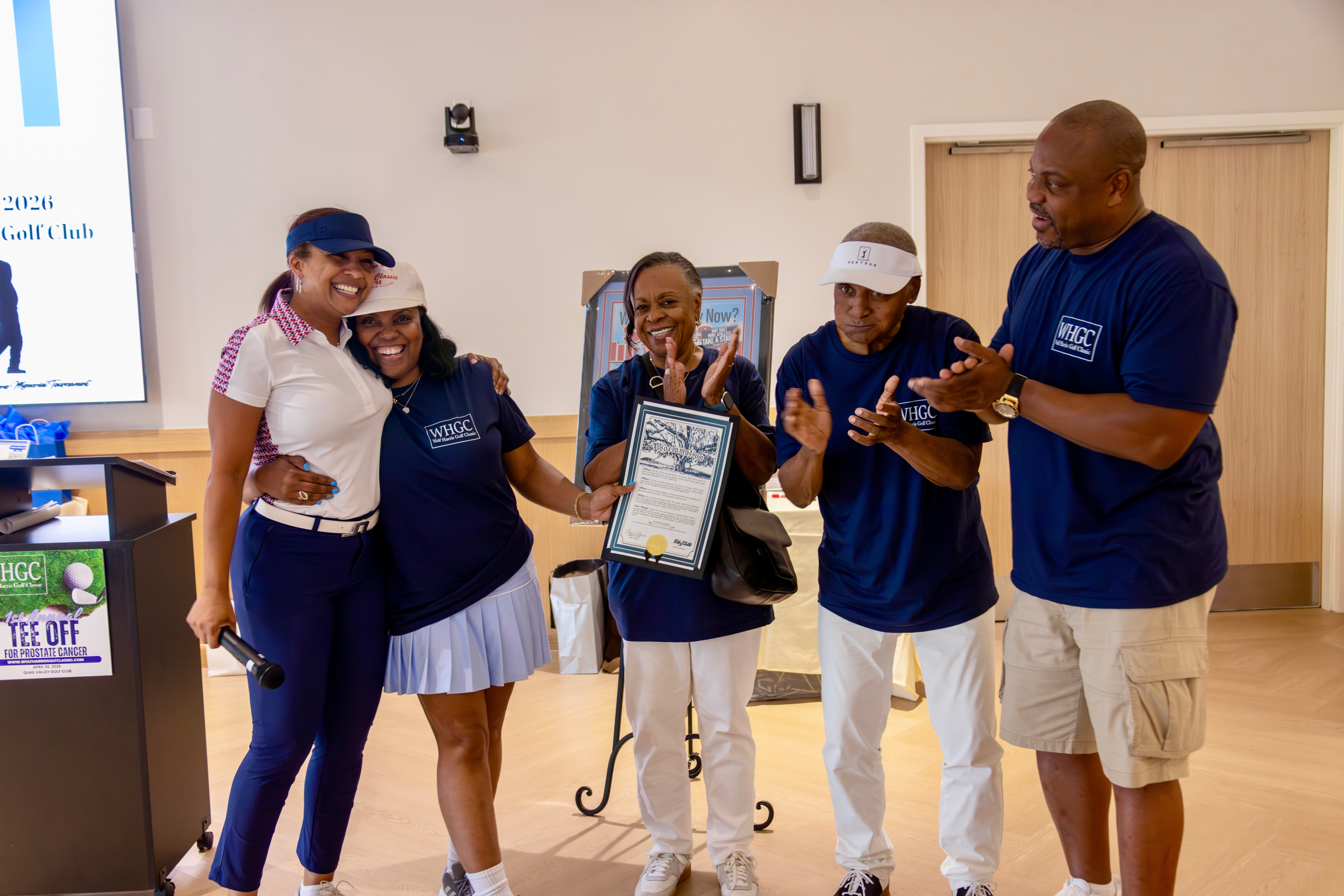 Group of five people standing indoors, smiling, with one holding a framed certificate, engaging in conversation.