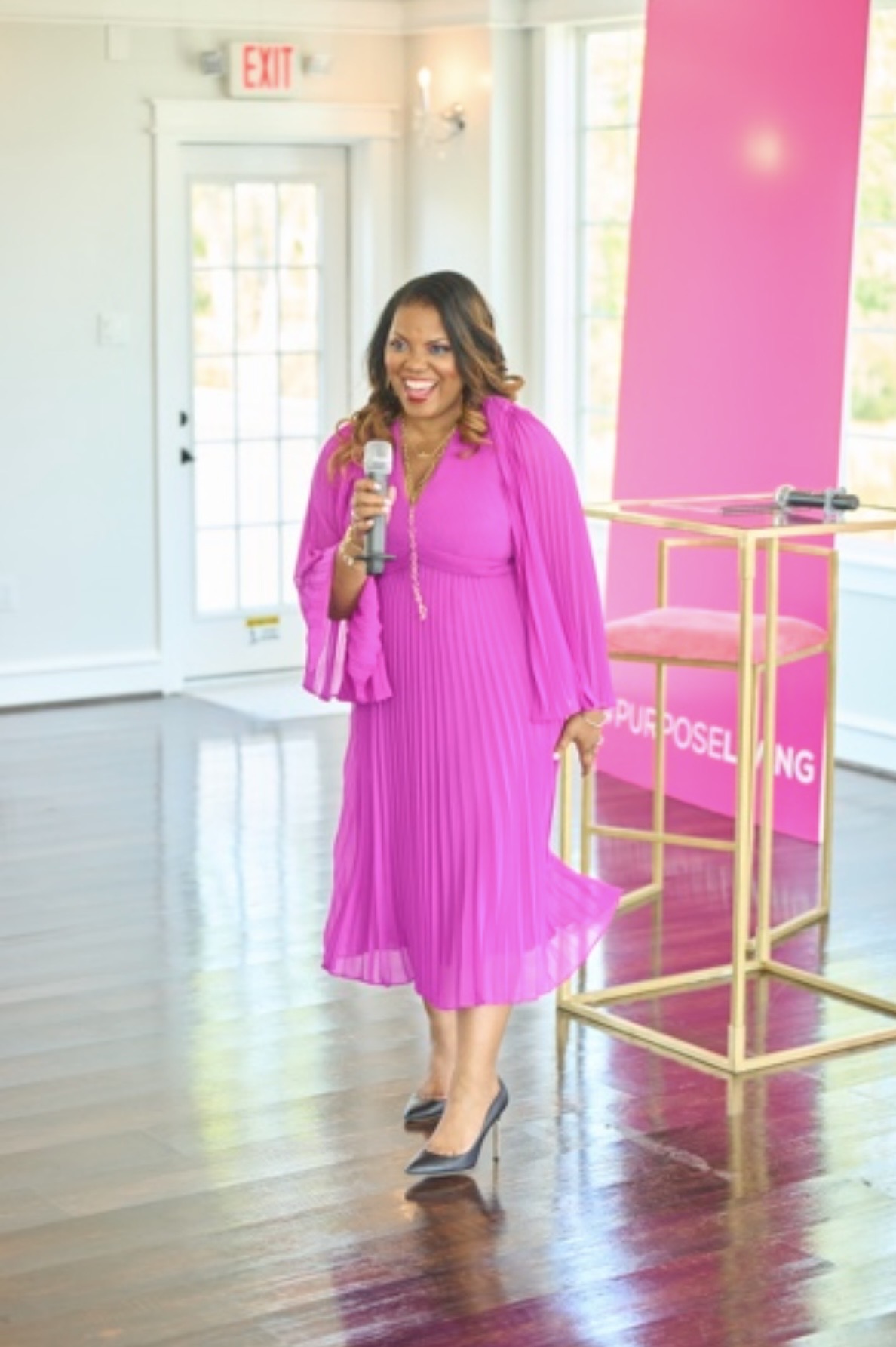 Woman in pink dress and blazer holding a microphone, smiling, standing indoors near a pink display and gold railing.