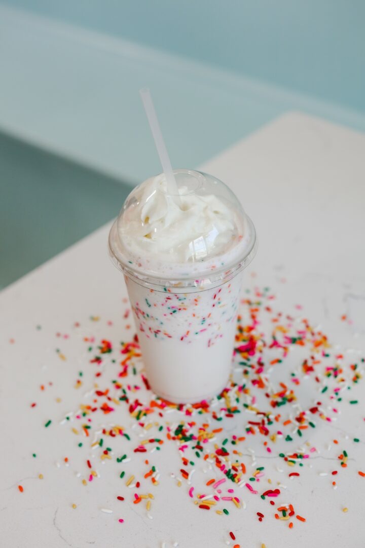 Milkshake with whipped cream and a straw in a clear cup, surrounded by colorful sprinkles on a white surface.