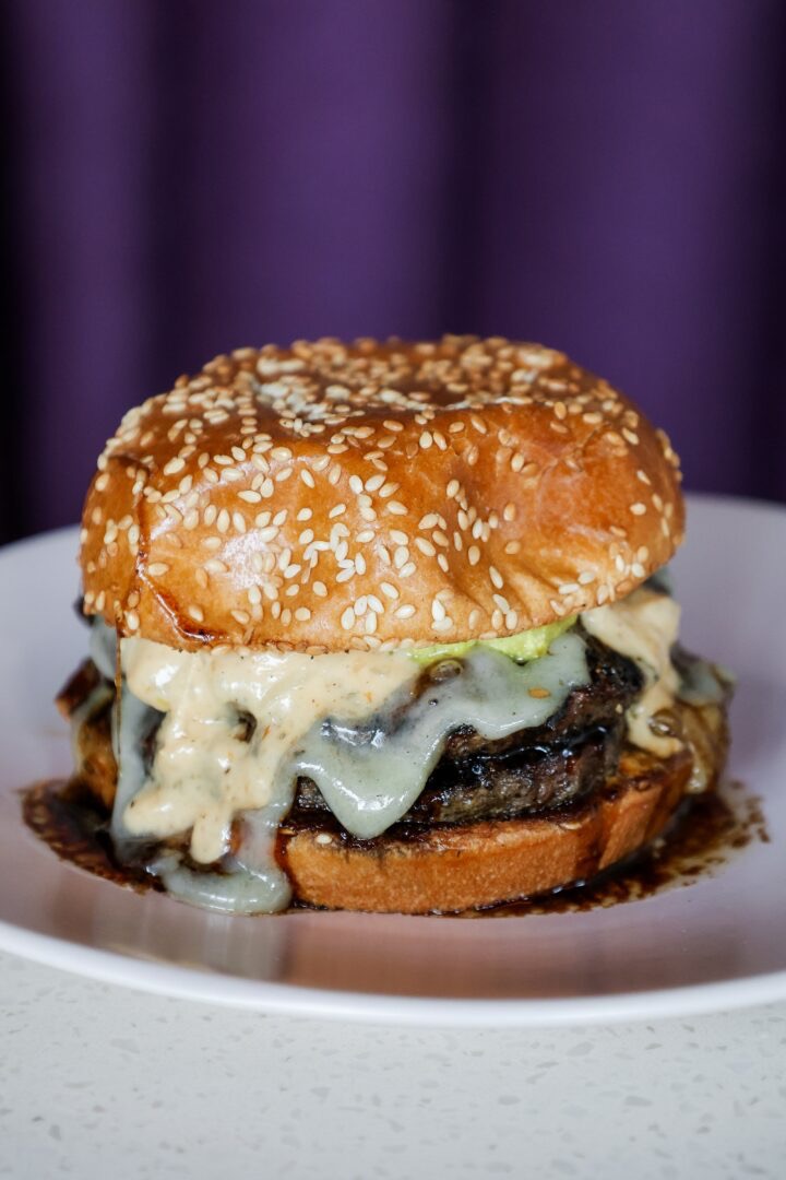 Cheeseburger with sesame seed bun, melted cheese, and beef patty on a white plate, purple background