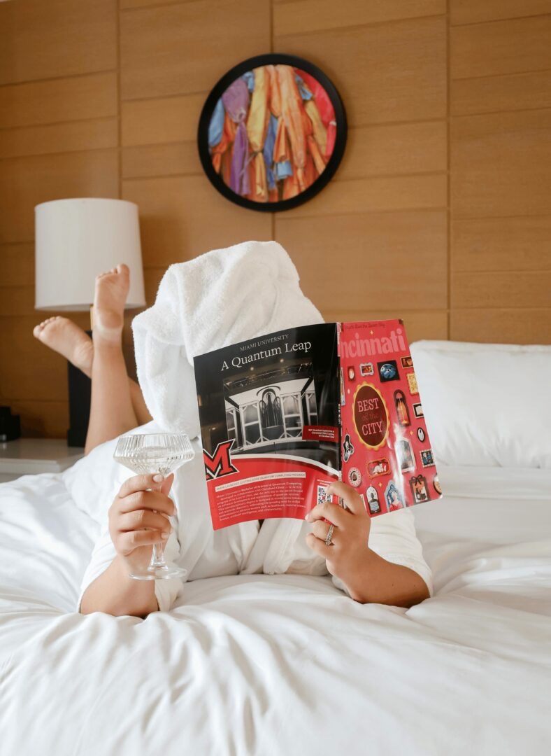 Person lying on bed reading a book, with legs raised, in a room with wooden wall and colorful artwork, white bedding.