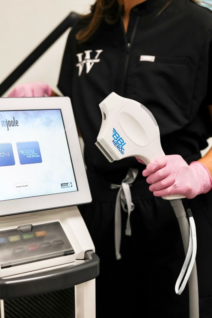 Person in black uniform holding a white medical device with pink gloves, next to a screen displaying blue icons.