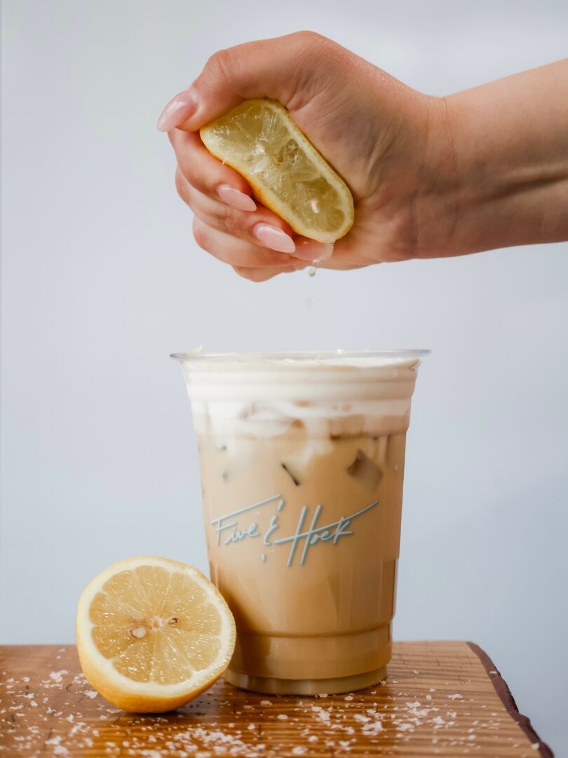 Hand squeezing lemon over iced coffee on wooden surface with lemon half nearby, against plain background.