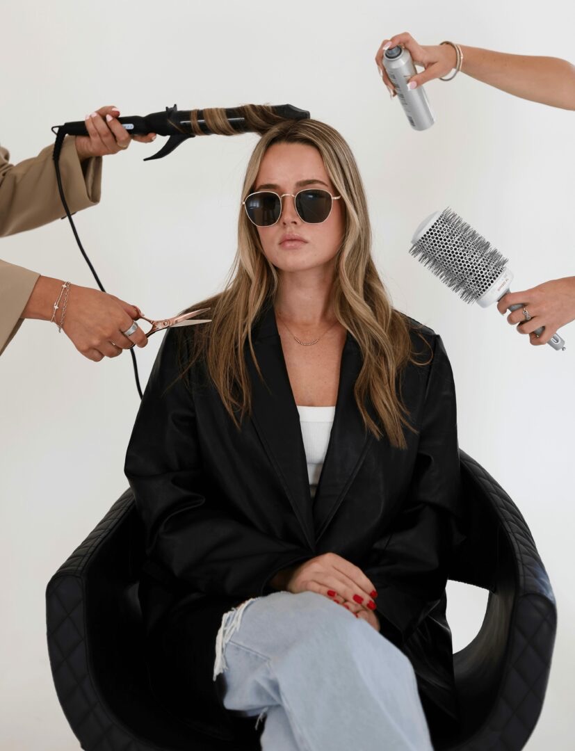 Woman with sunglasses sits calmly as hairstyling tools and spray are used around her.