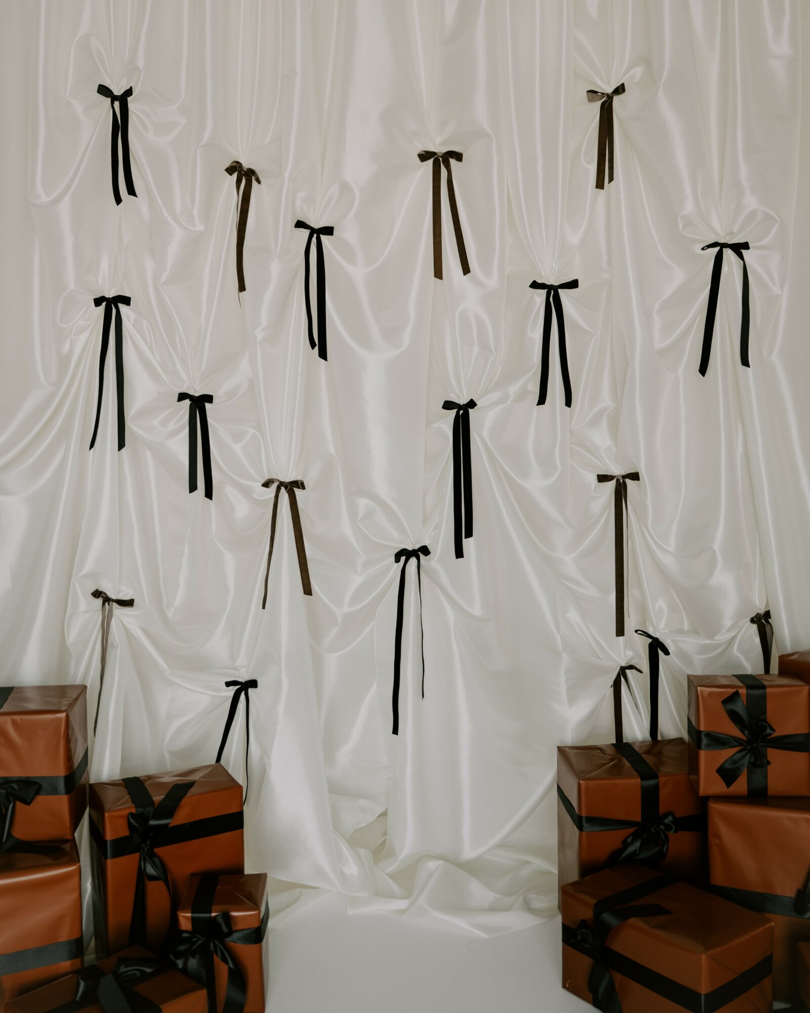 White curtain backdrop with black and brown bows, surrounded by wrapped gift boxes with black ribbons, in a festive setting.
