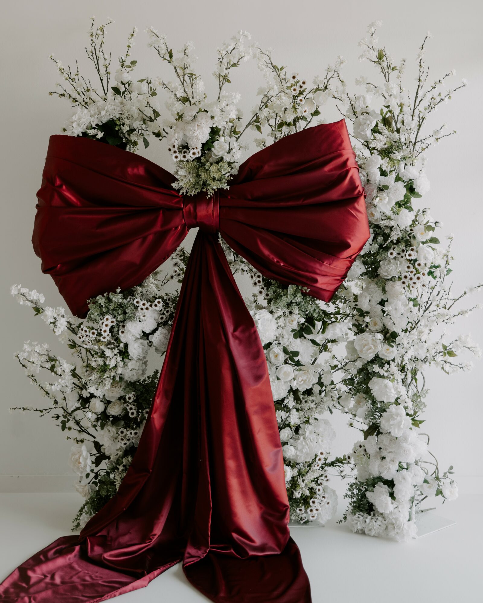 Decorative arrangement with white flowers and a large red satin bow in front, against a plain background.