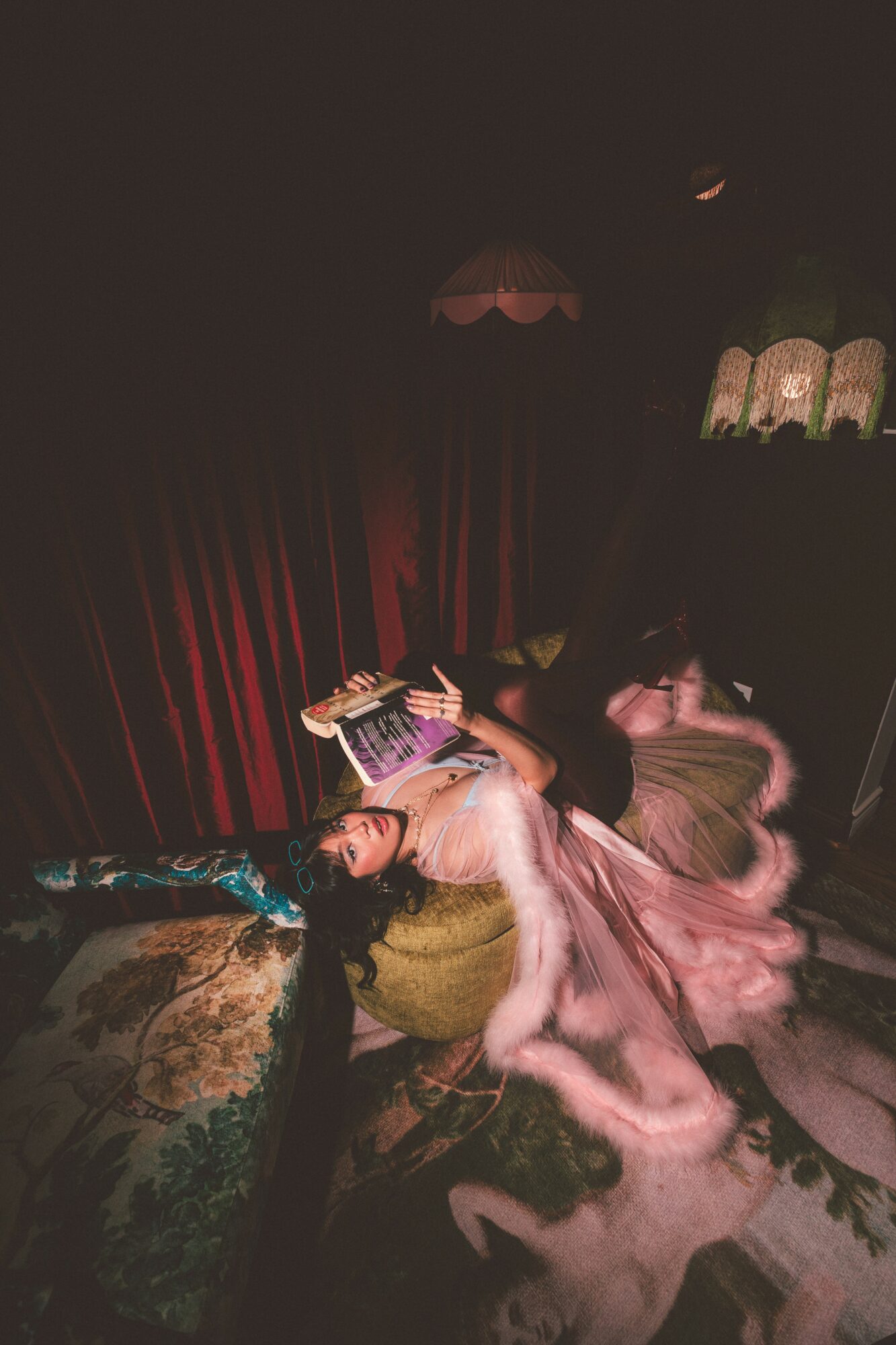 Person lying on a sofa reading a book, with a pink dress and long hair, in a dimly lit room with red curtains.