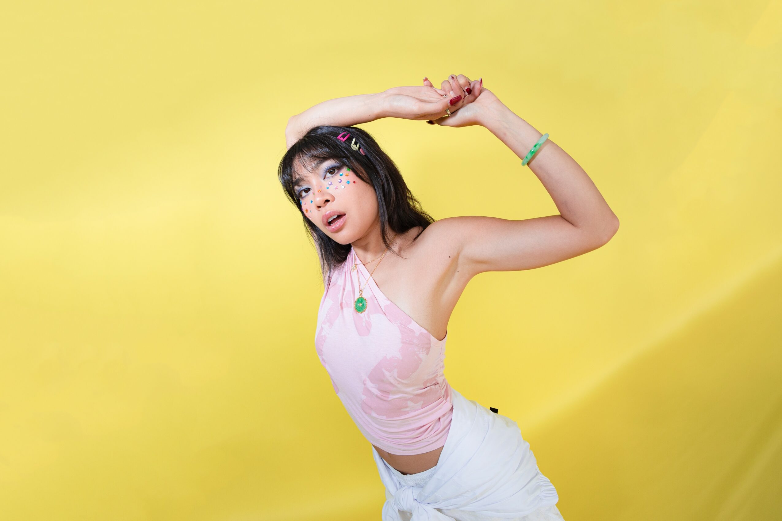 Young girl with dark hair and bangs, wearing a pink top and white pants, poses with arms above her head against yellow background.