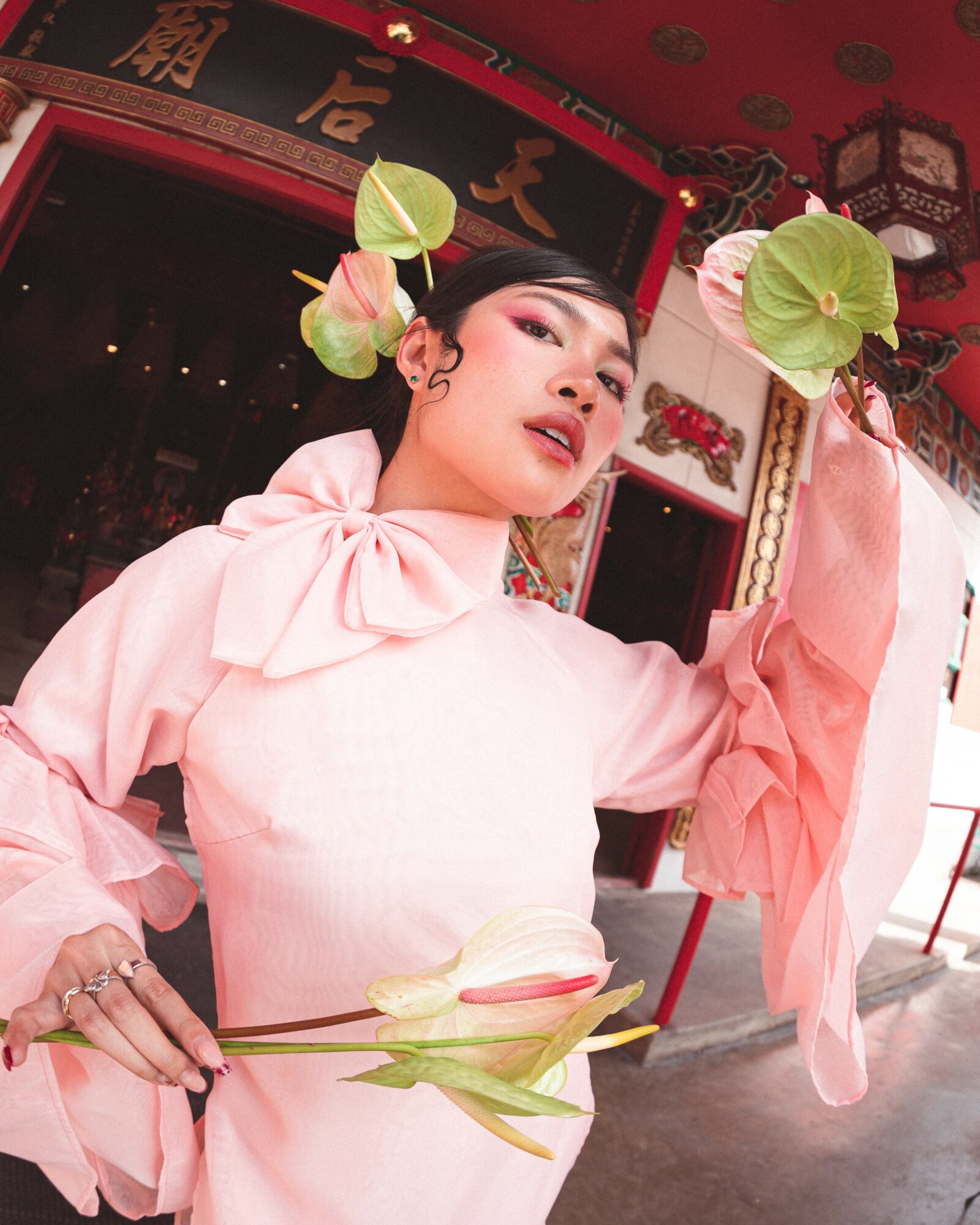 Woman in pink traditional dress with flowers in hair, holding a flower, in a decorated interior with red and gold accents.