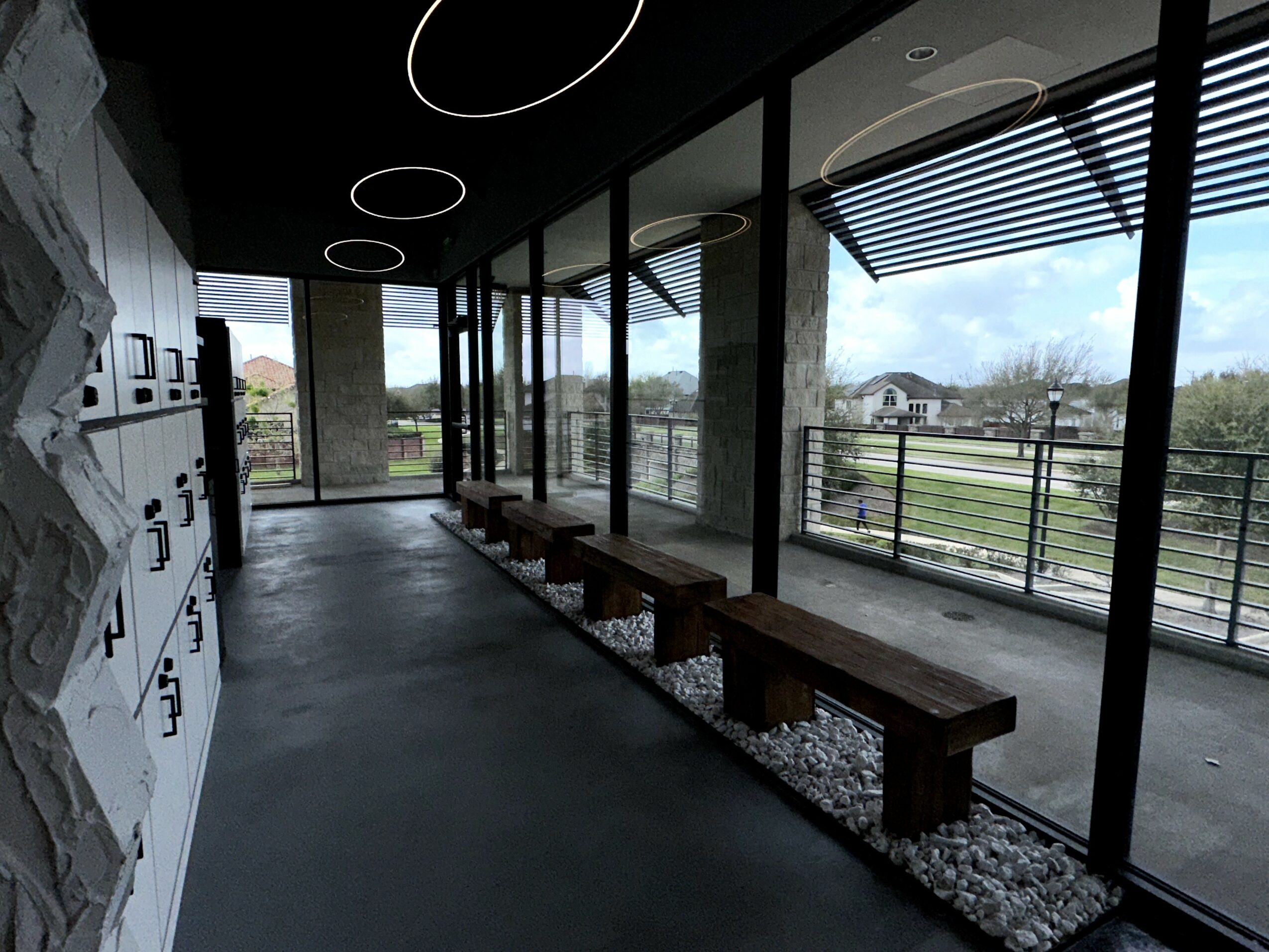 Indoor corridor with large glass windows, wooden benches, and modern ceiling lights, overlooking an outdoor landscape with fields and trees.