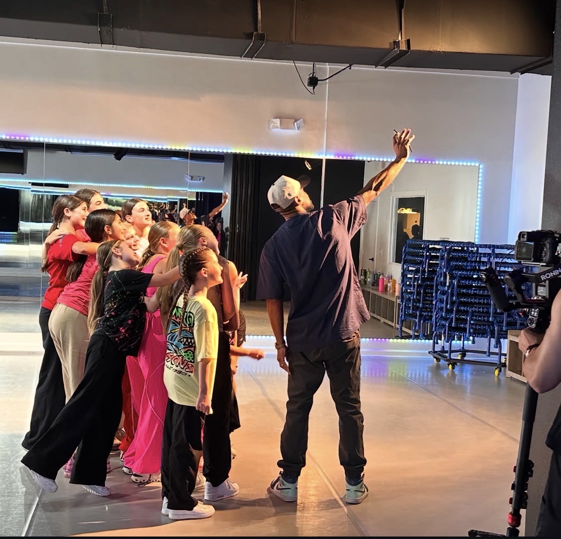Group of children watching a person with white hair raise an arm in a dance studio with mirrors and stacked chairs.