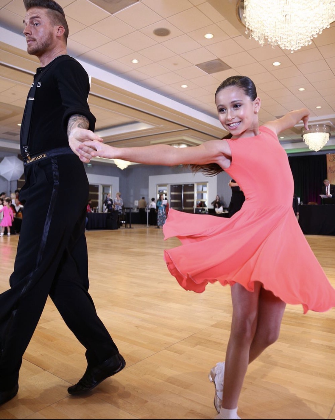 A young woman in a pink dress and a young man in black dance together in a ballroom, smiling and holding hands.